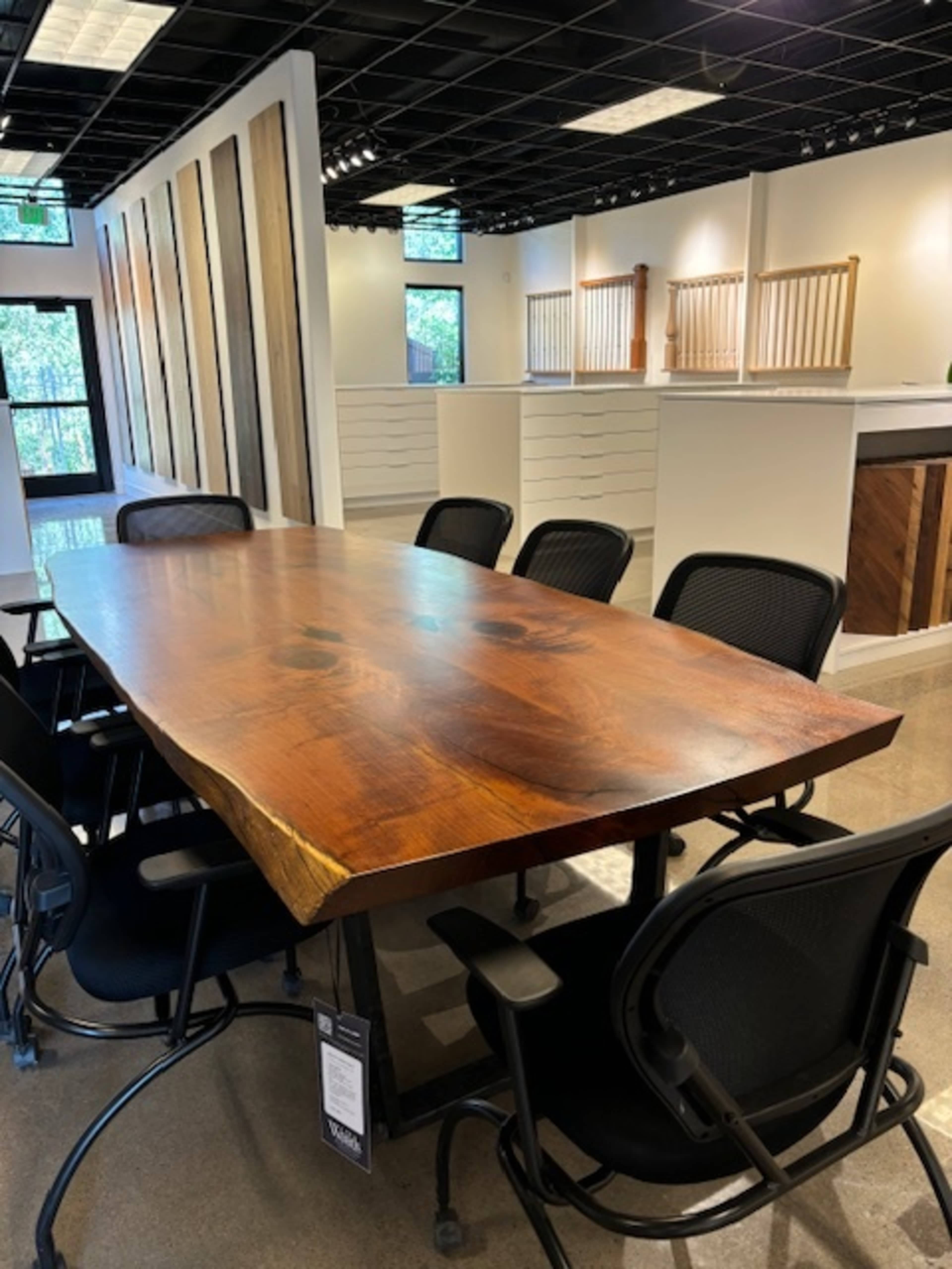 A large wooden conference table with a natural edge is surrounded by several black office chairs in a brightly lit room with wooden accent walls.