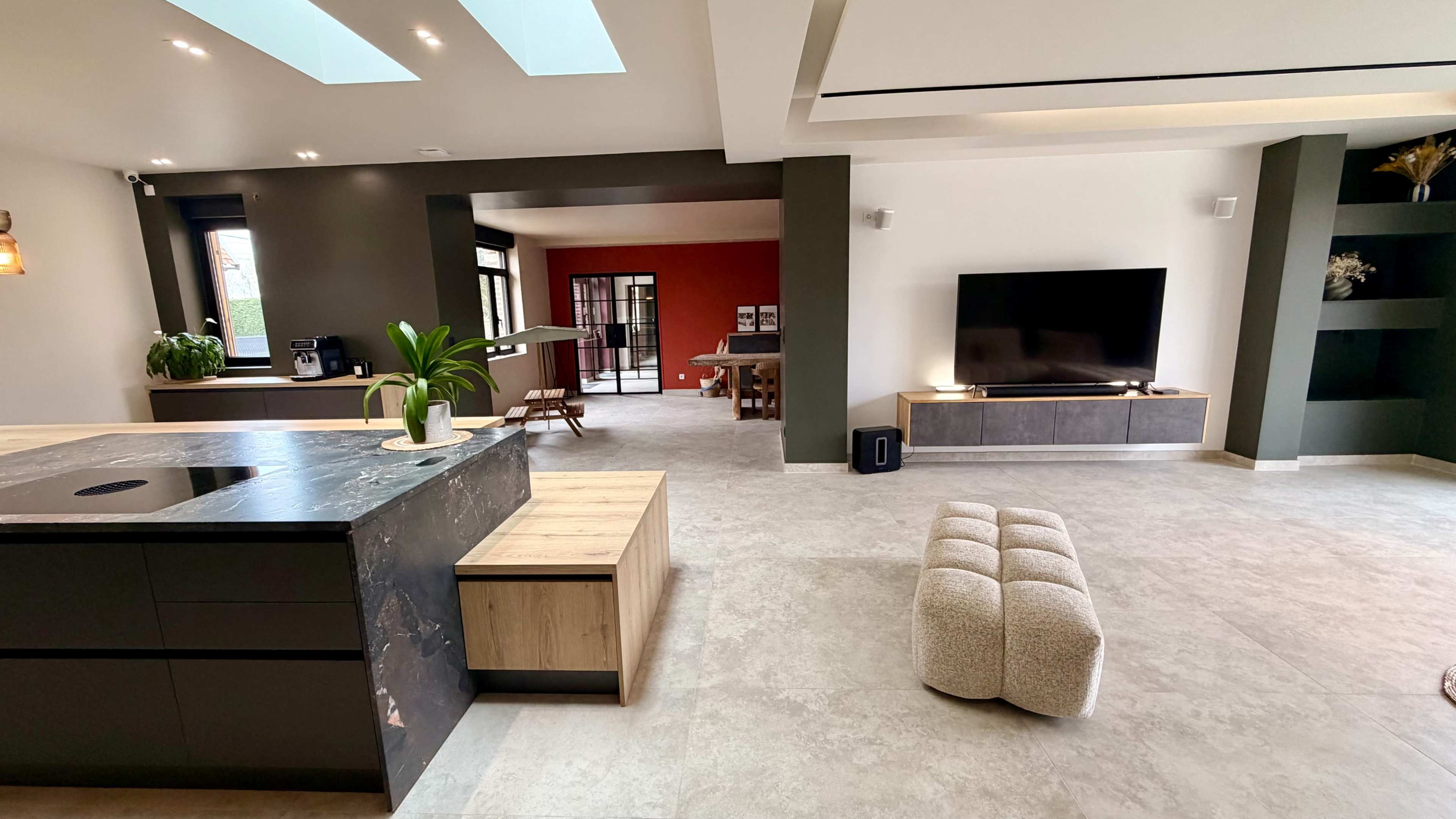 The image shows a modern interior space featuring an open-concept living area with a kitchen, dining area, and a living room equipped with a large television.