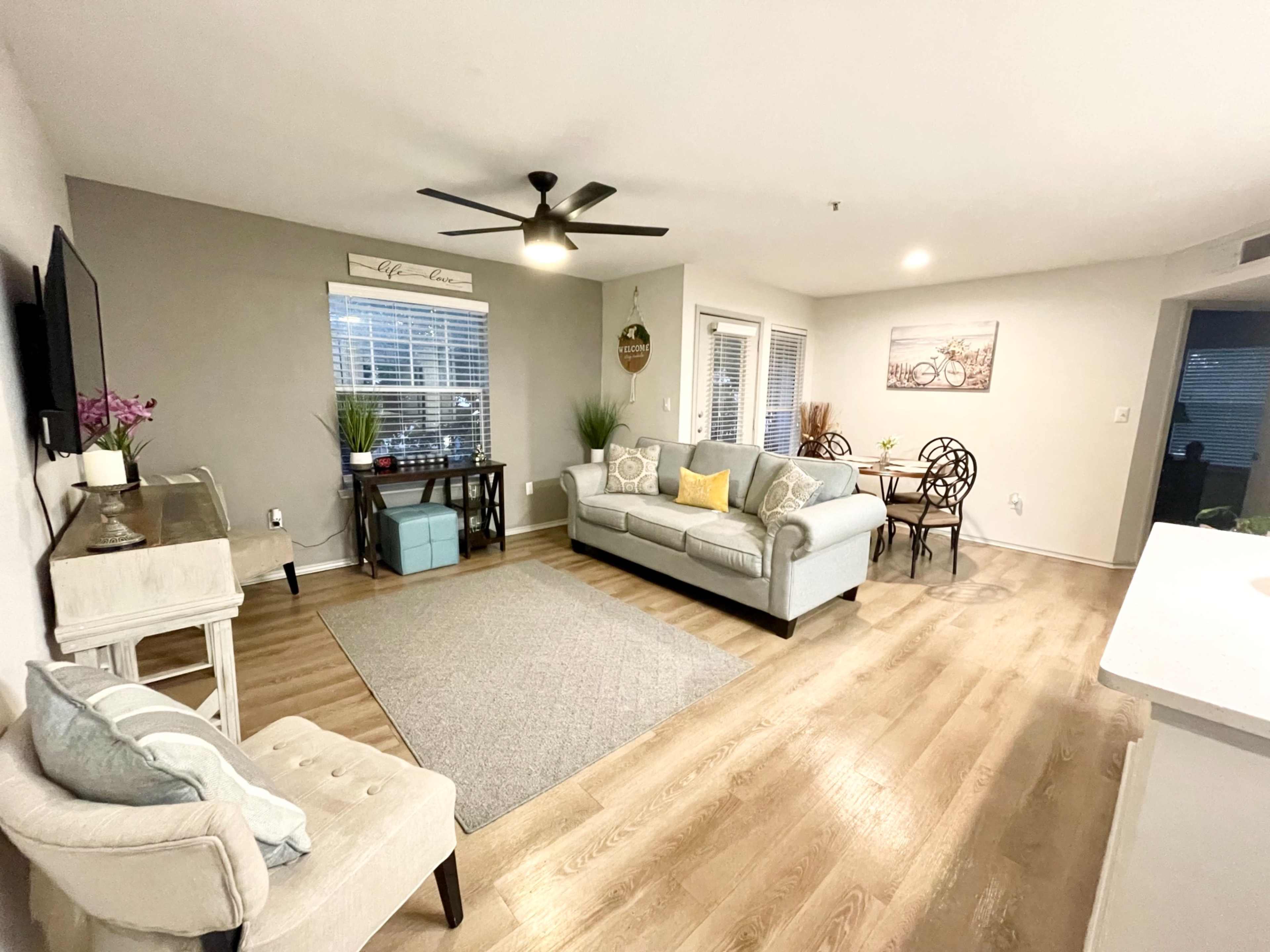 A modern living room with a sofa, armchair, coffee table, and a dining area, featuring a light color scheme and wooden flooring.