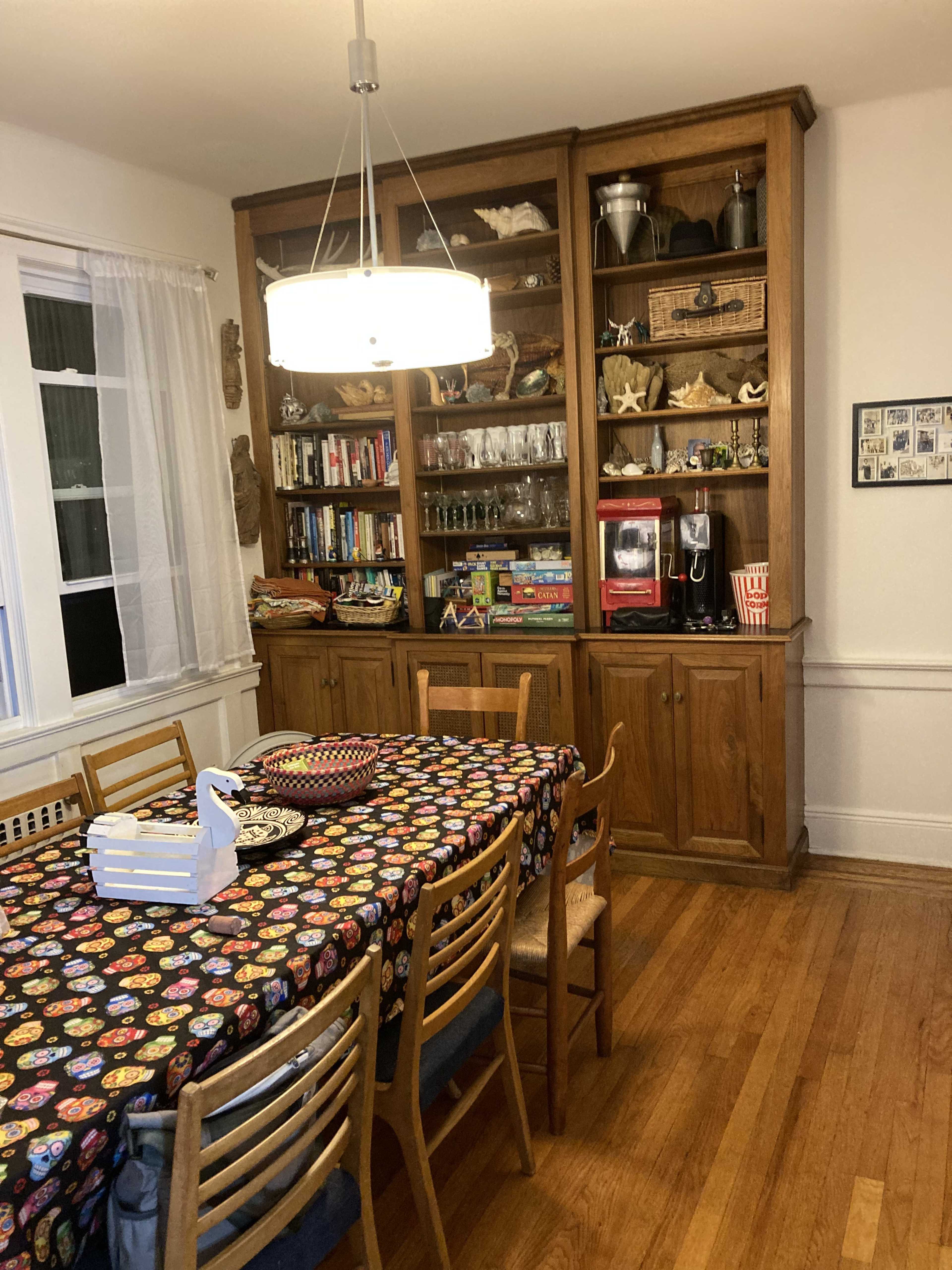 A dining room features a table set with a colorful tablecloth and surrounded by wooden chairs, while a large bookshelf displays various items and games.