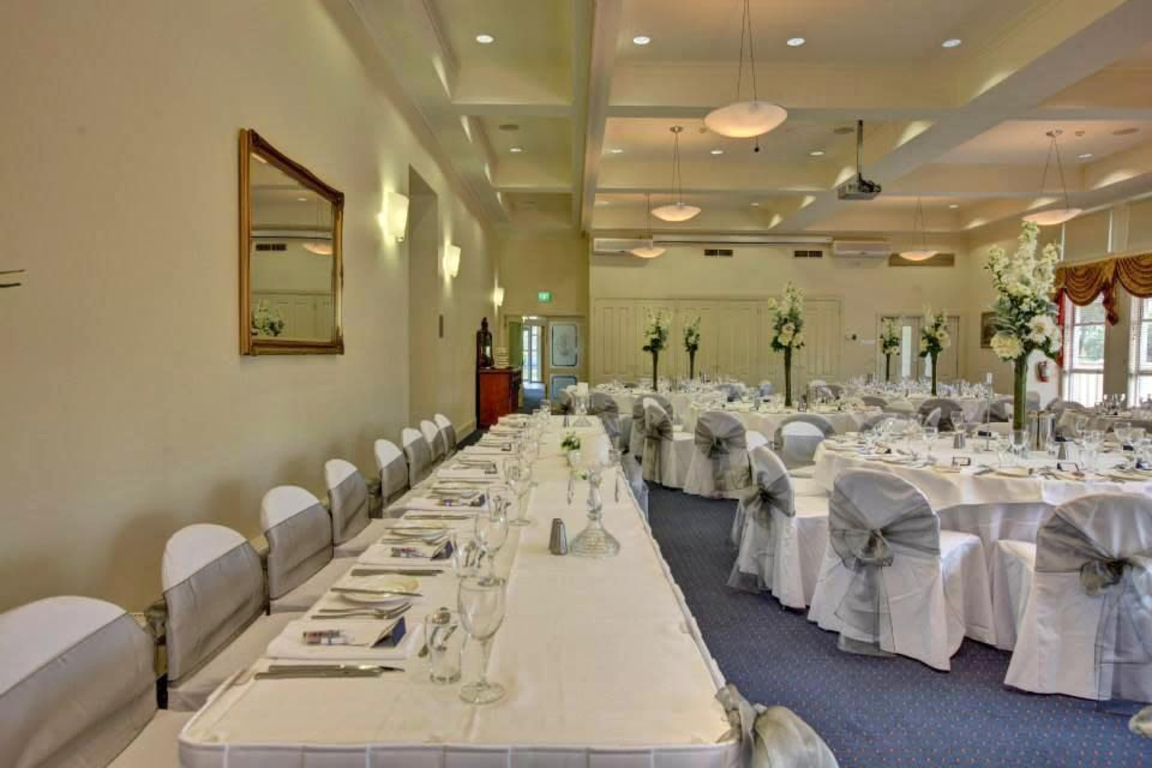 A spacious banquet hall is set with elegantly arranged tables covered in white cloths and adorned with floral centerpieces.