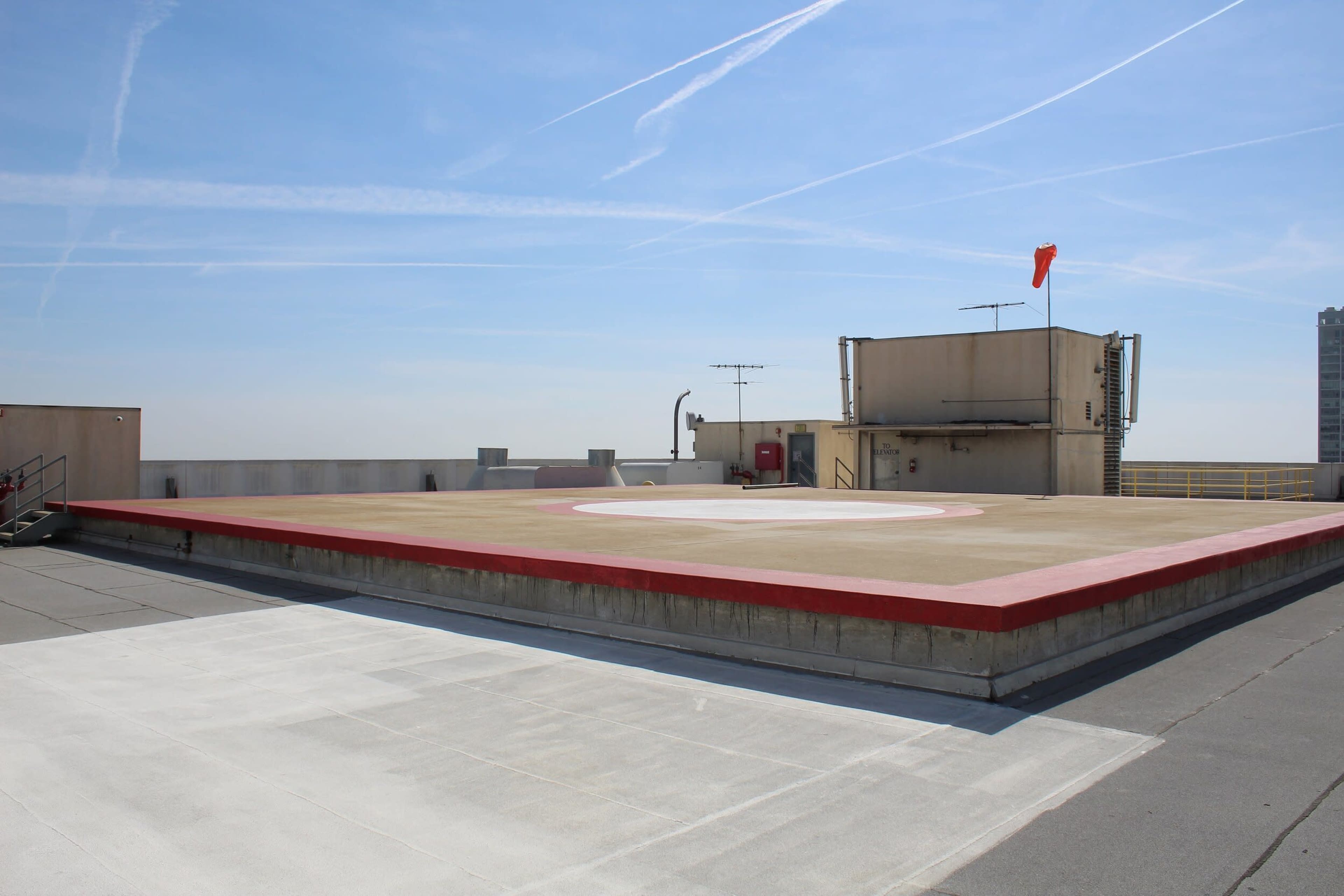 A flat rooftop helipad with a circular marking in the center and a red border, surrounded by a clear blue sky with some contrails.
