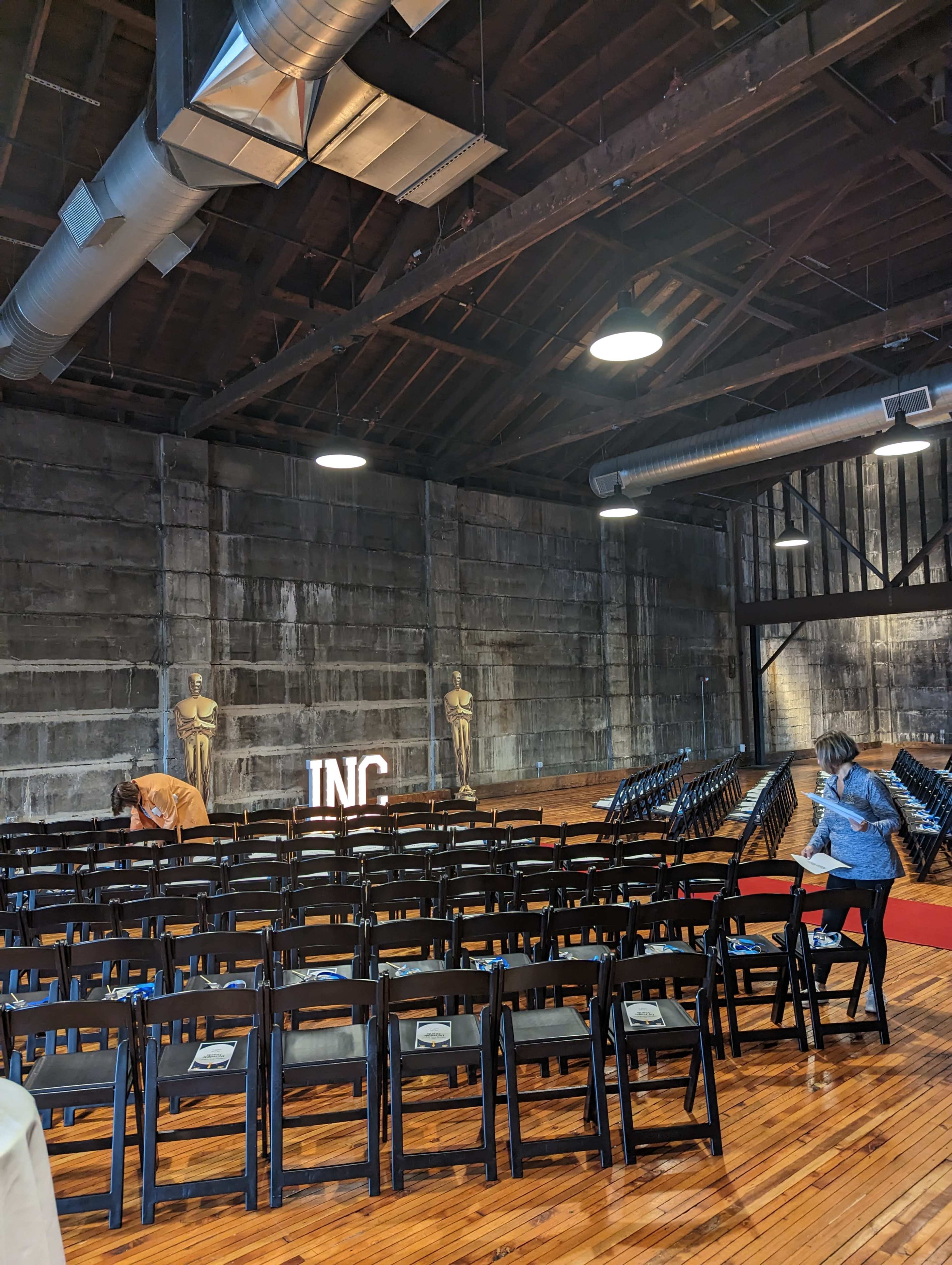 An indoor event space with rows of black chairs set up facing a lit sign that says "INC," and a few people preparing the area.