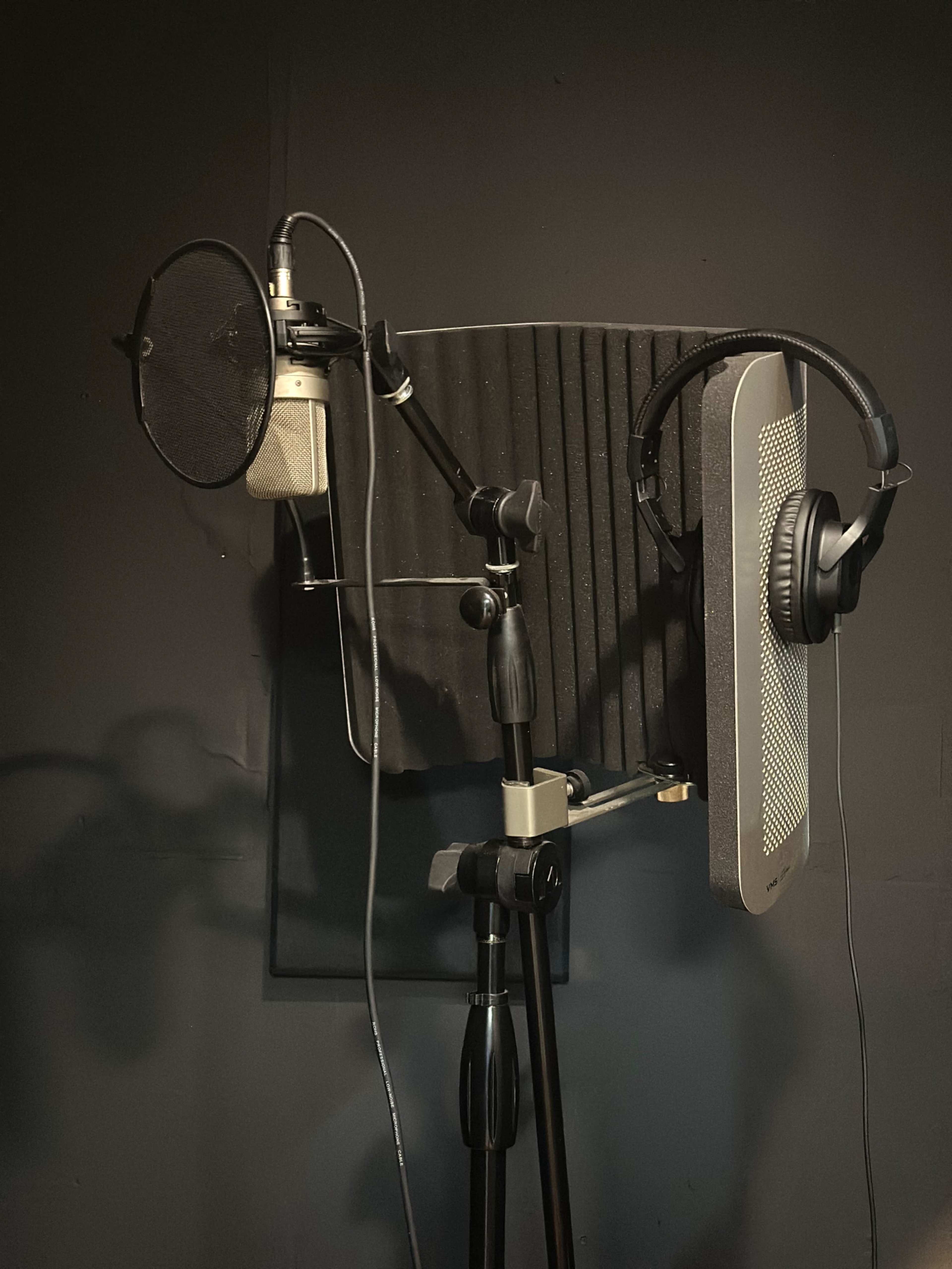 The image shows a microphone setup with a pop filter, sound panel, and headphones mounted on a stand against a dark background.