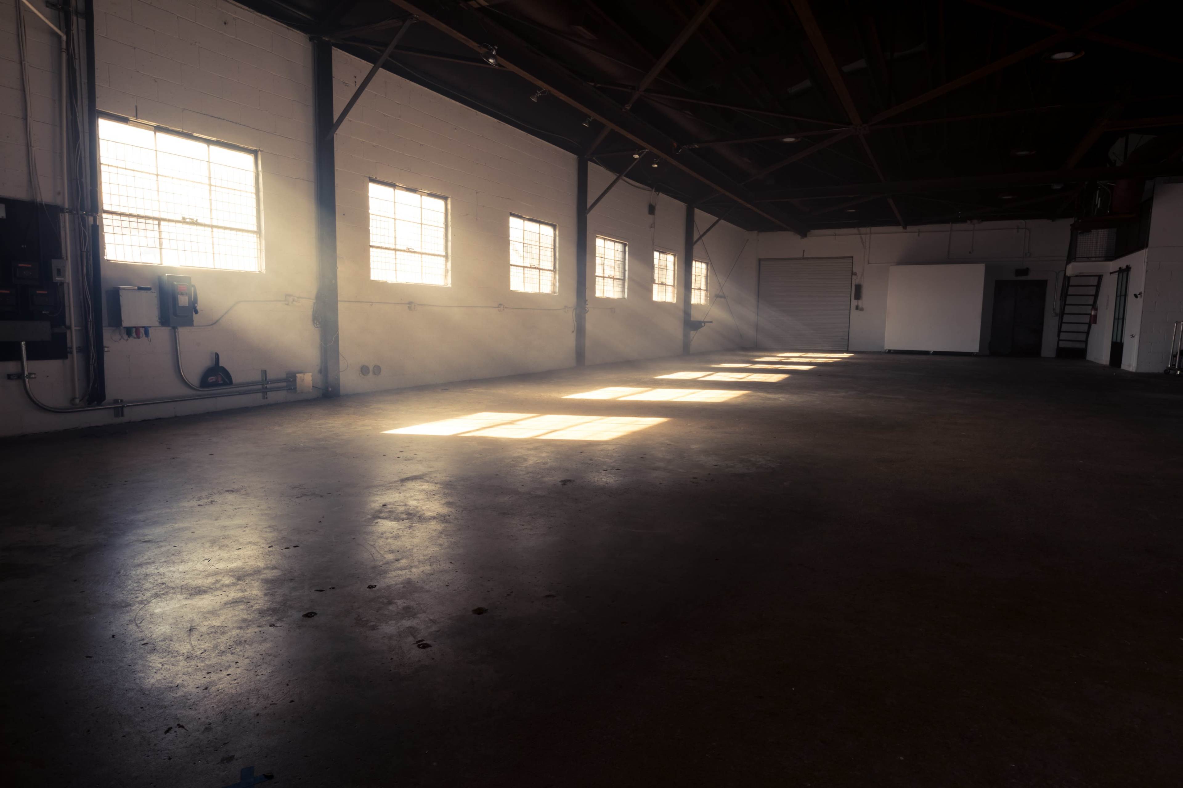 A sunlit industrial space features an empty concrete floor and large windows casting shadows along the walls.