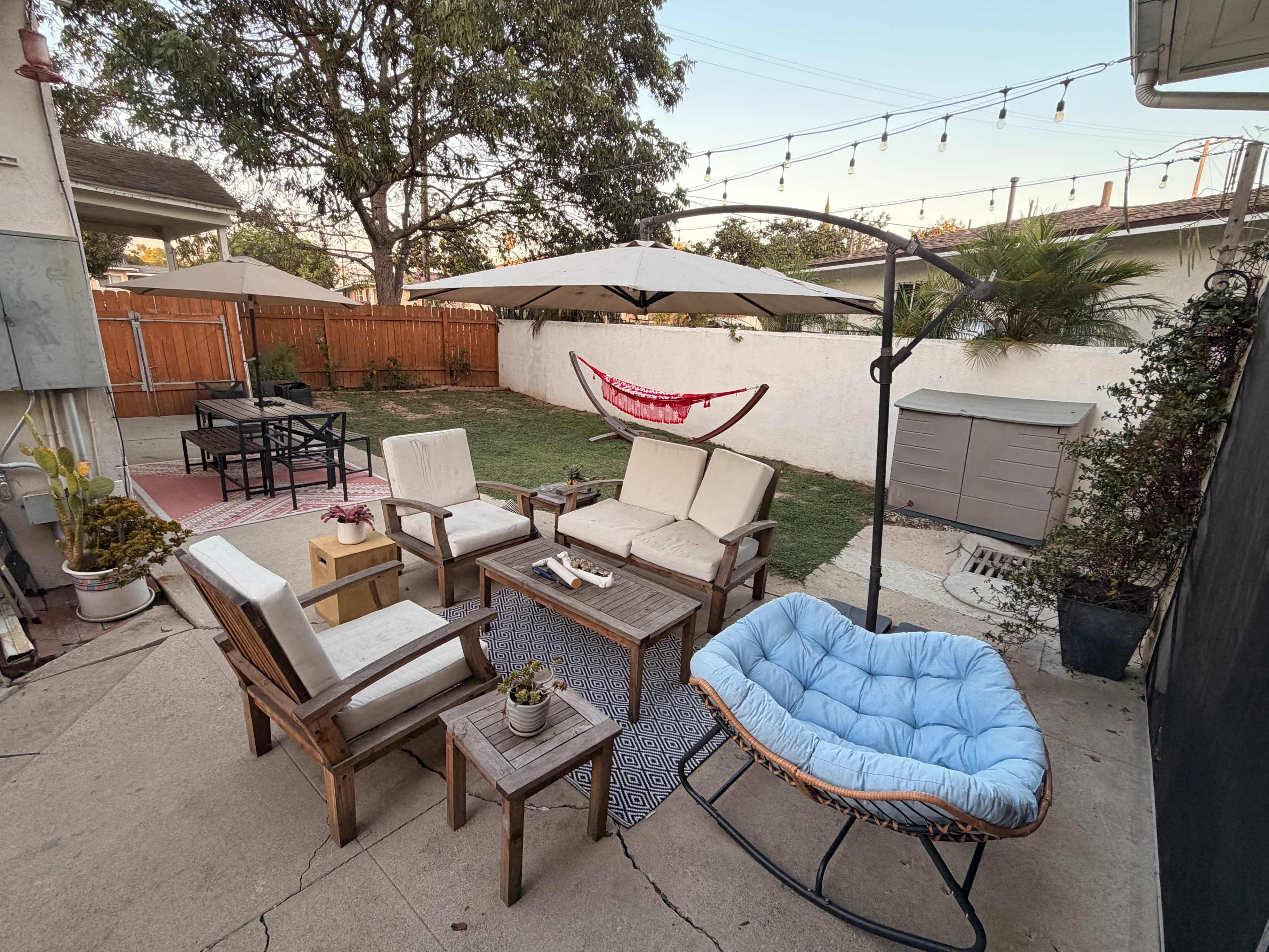 The image shows a backyard seating area with wooden furniture, a hammock, and an umbrella over a circular swing, surrounded by a garden space.