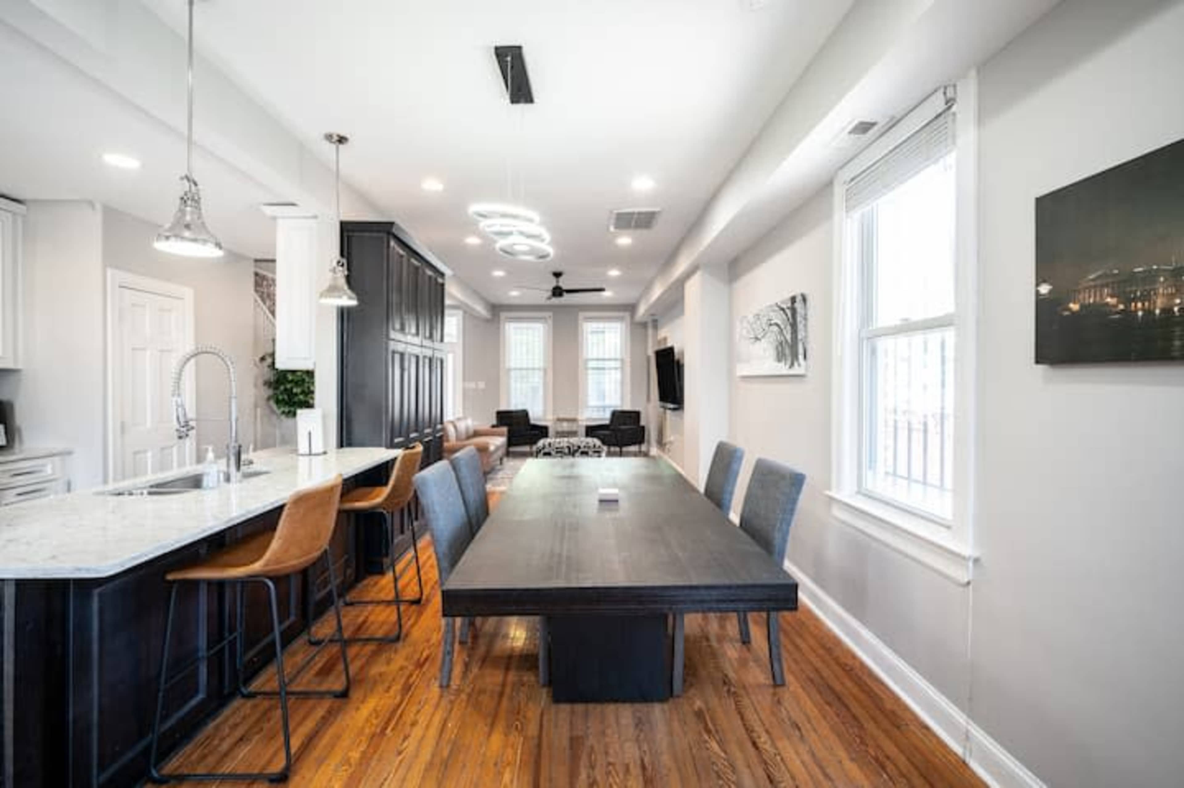 The image shows a modern kitchen and dining area featuring a long black dining table, bar stools at the counter, and a mix of seating arrangements along with large windows letting in natural light.