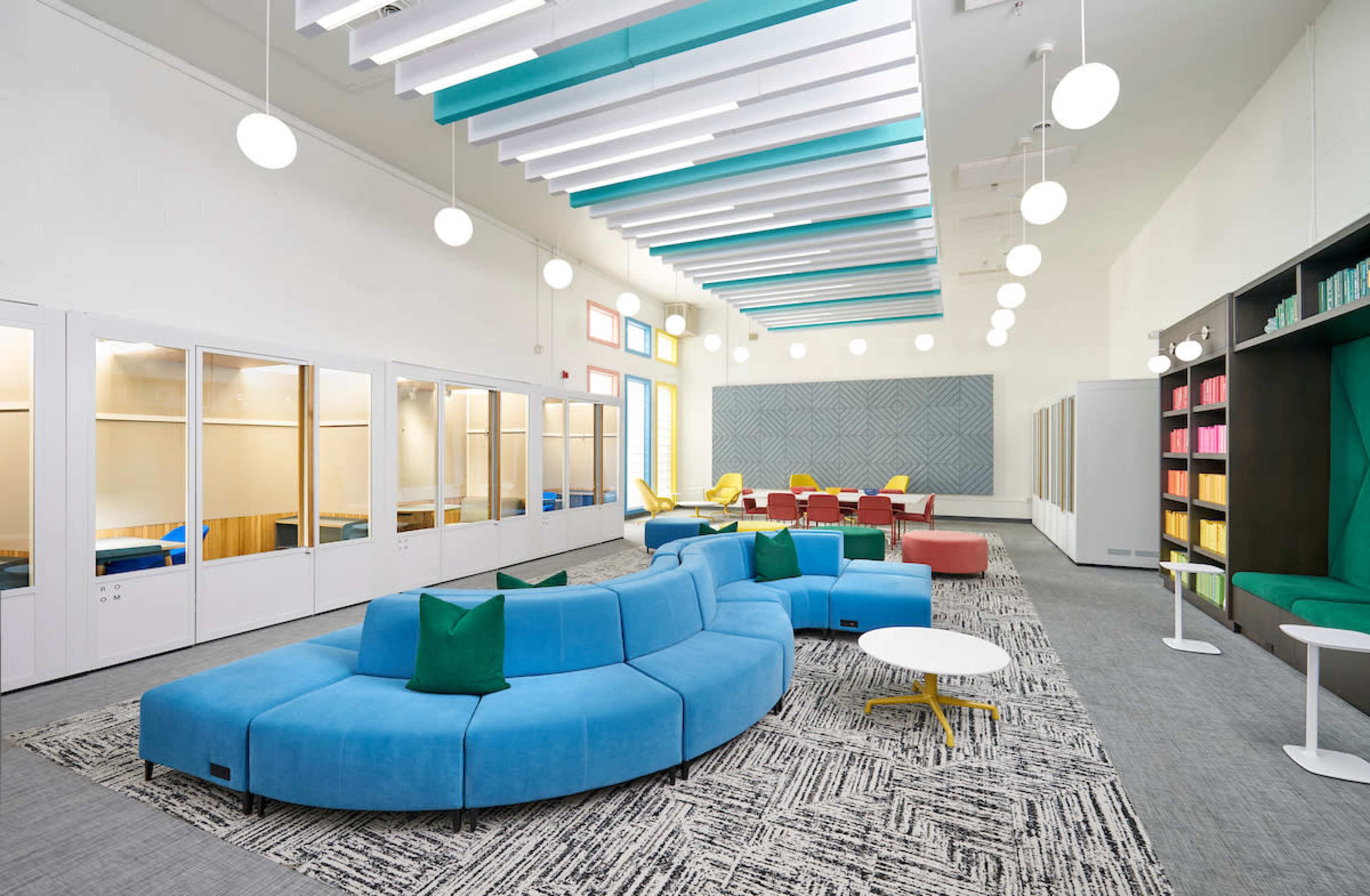 A modern, brightly lit room featuring a blue sectional sofa, various seating areas, and glass partitions for individual workspaces.
