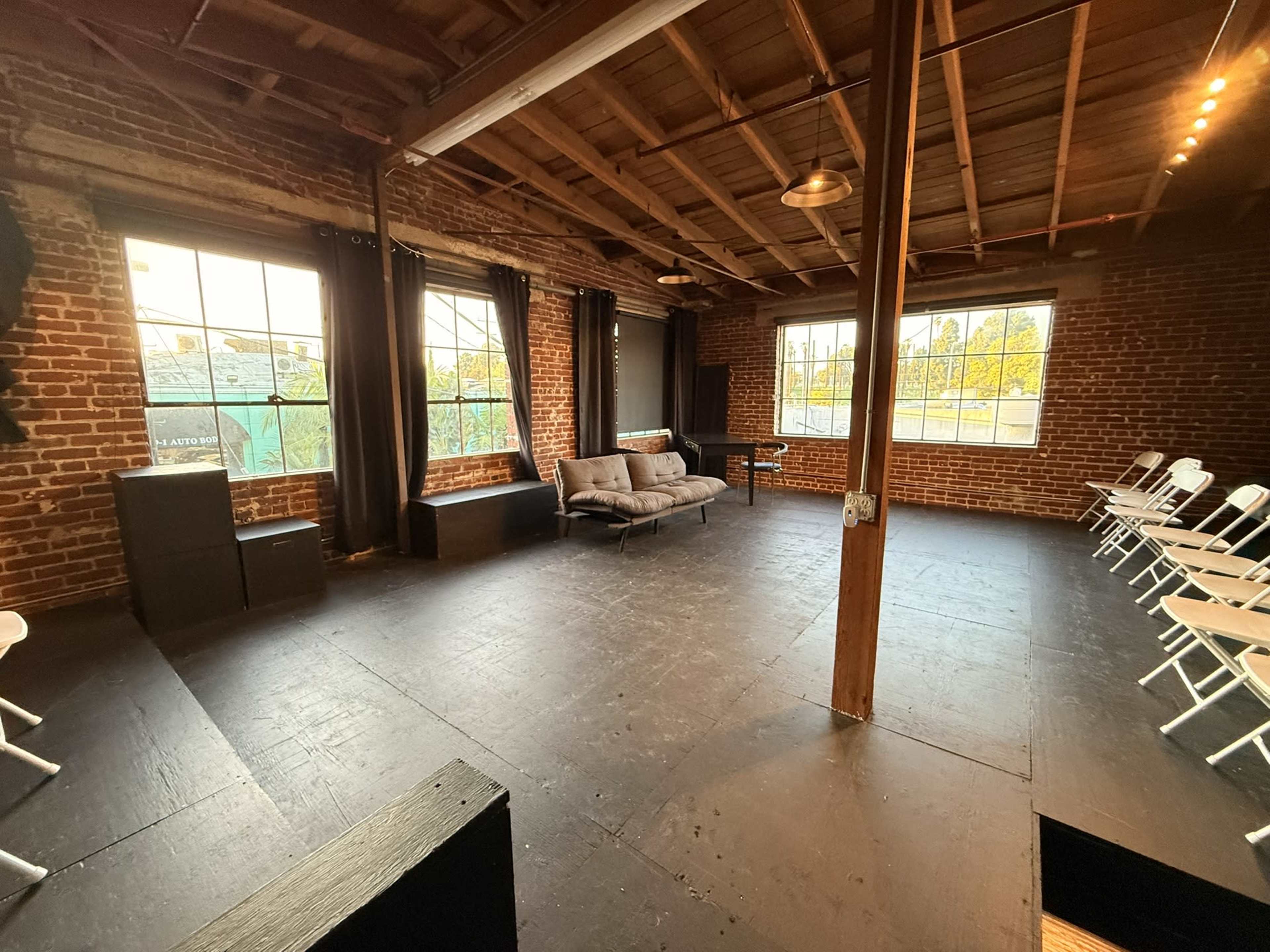The image shows a spacious room with exposed brick walls, large windows, and a mix of seating options including a sofa and white folding chairs.
