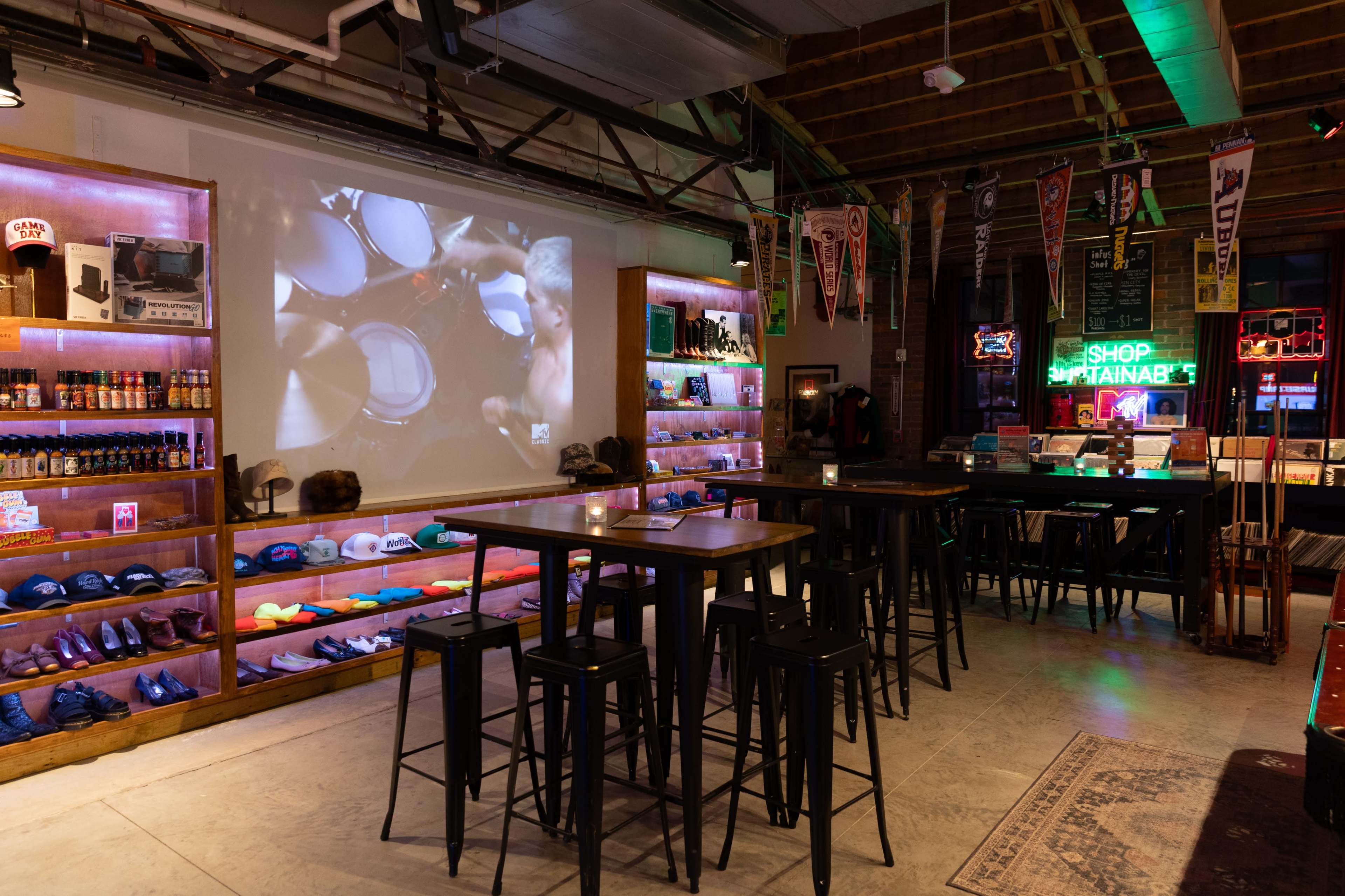 The image shows a lively bar area with tall tables and stools, decorated with colorful shelves displaying merchandise and a large screen showing a music performance in the background.
