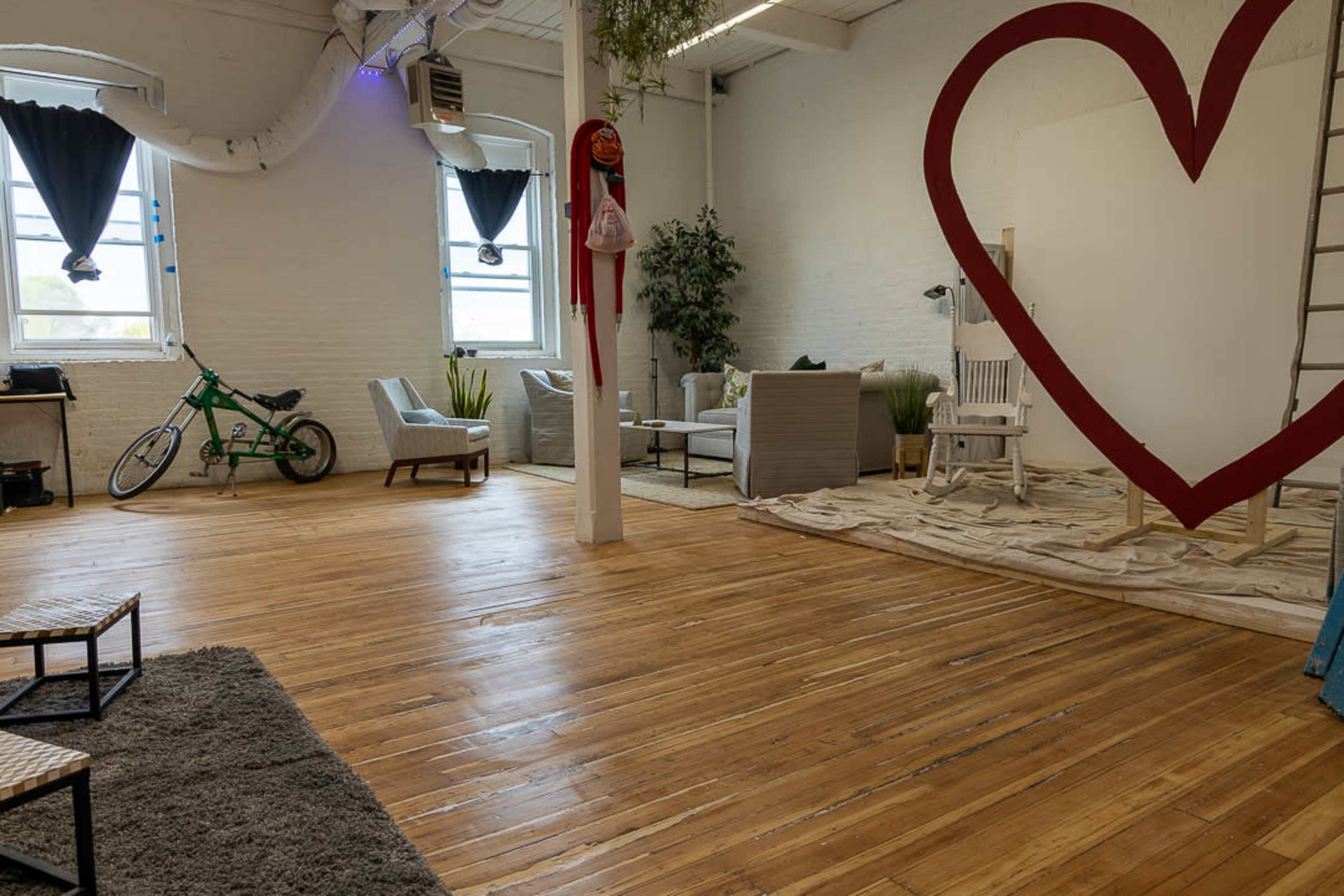 The image shows a spacious, well-lit room with wooden floors featuring a large red heart decoration on the wall, seating areas with chairs and a sofa, and a bicycle leaning against the wall.