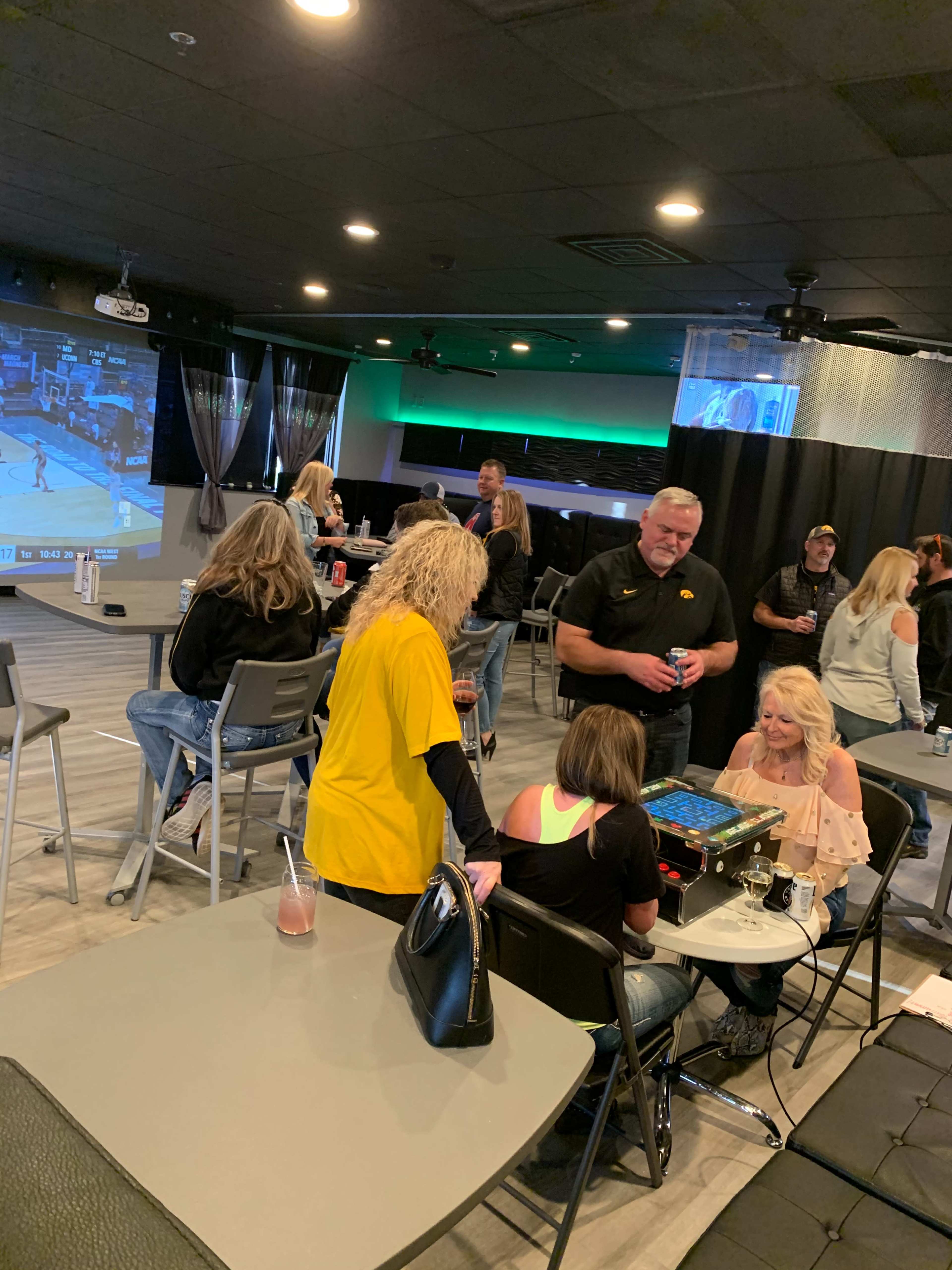 A group of people socializes in a casual setting with a large screen displaying a basketball game.