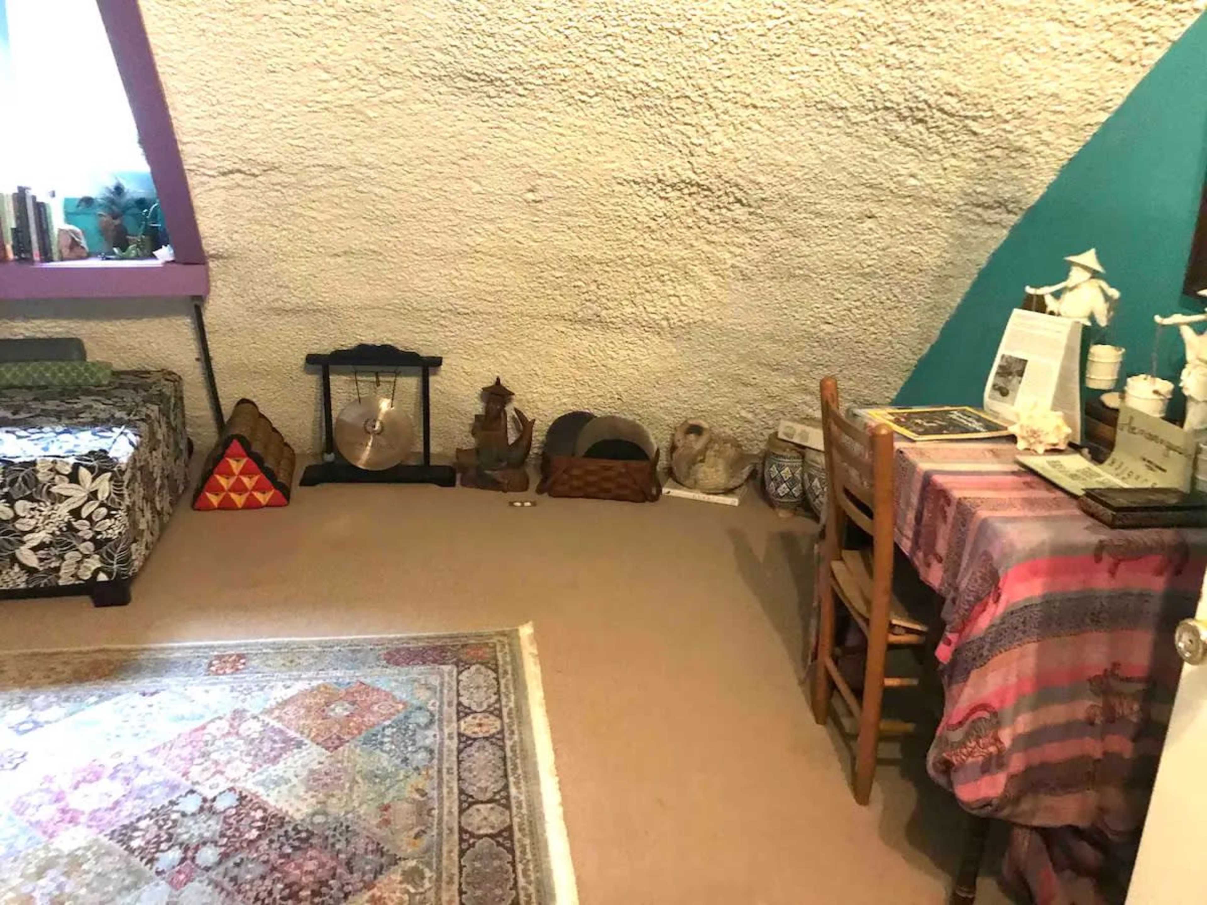 The image shows a cozy room with a textured white ceiling, a patterned rug on the floor, a wooden table with various items, and seating along the walls.