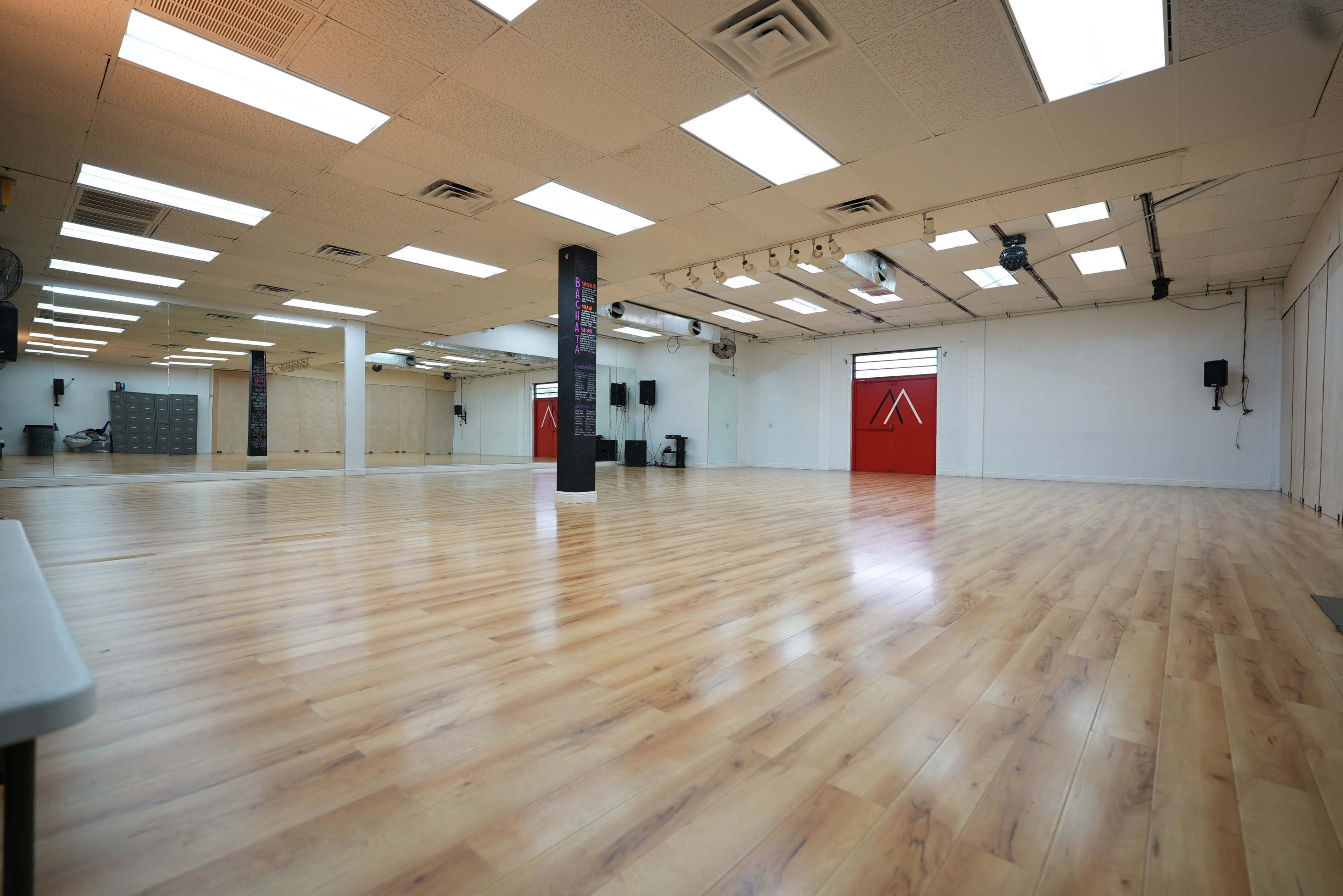 The image shows a large dance studio featuring a polished wooden floor, bright fluorescent lighting, and a red door on one wall.