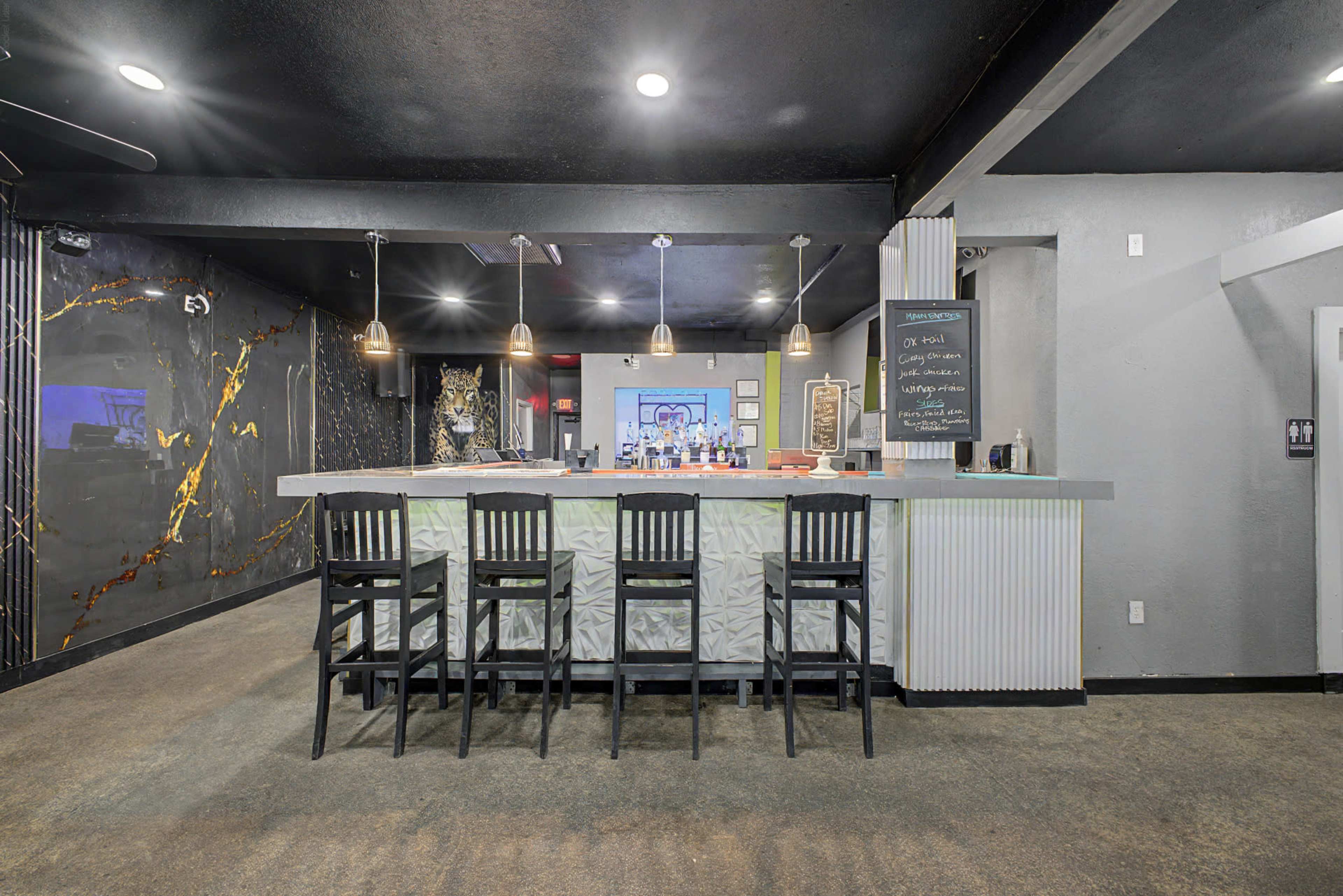 The image shows a modern bar area with a sleek counter, high stools, and decorative walls featuring dark colors and artistic elements.