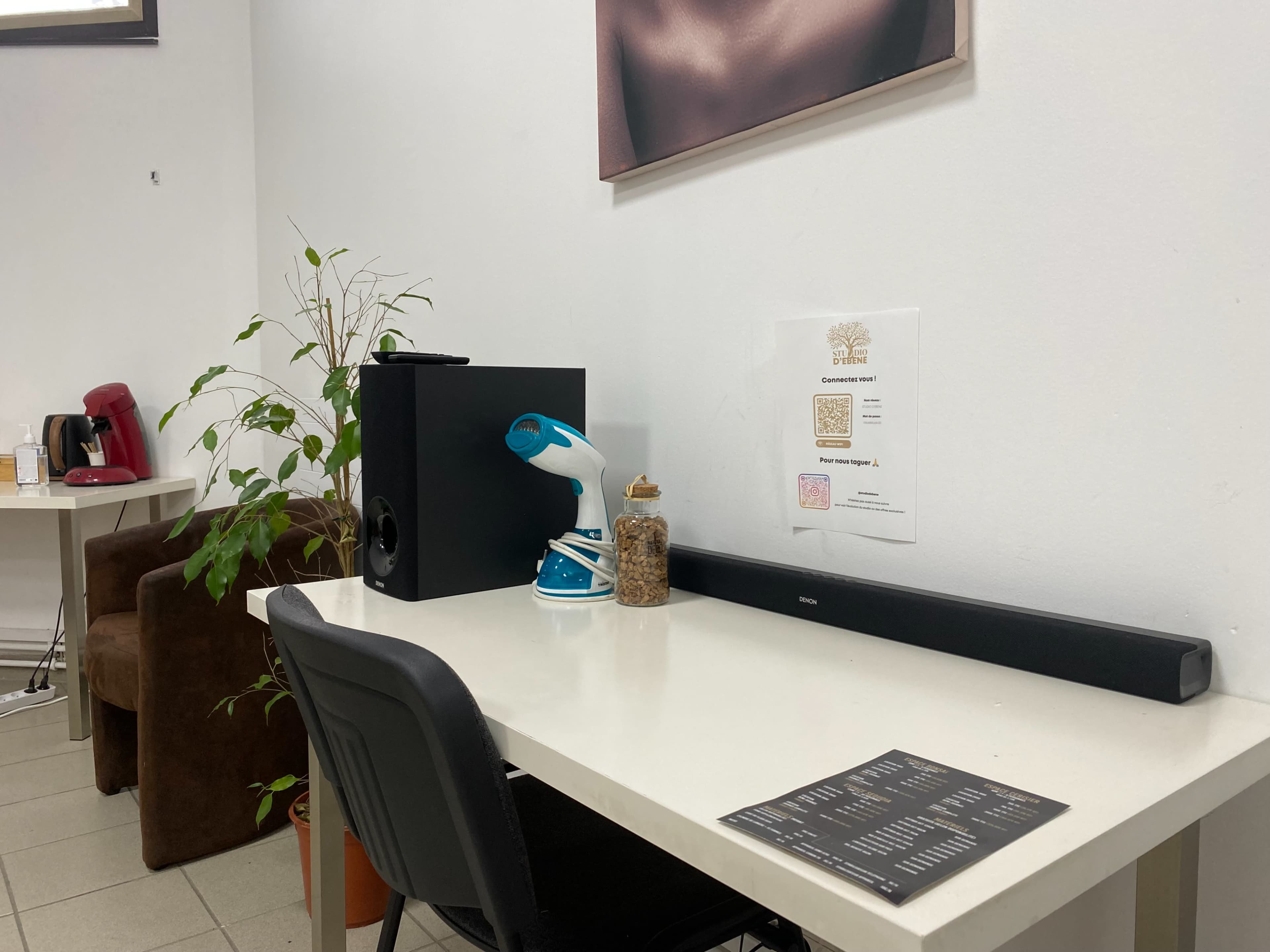 A small workspace features a white table with a black speaker, a spray bottle, a jar of items, and a printed QR code on the wall, alongside a plant and a brown chair.