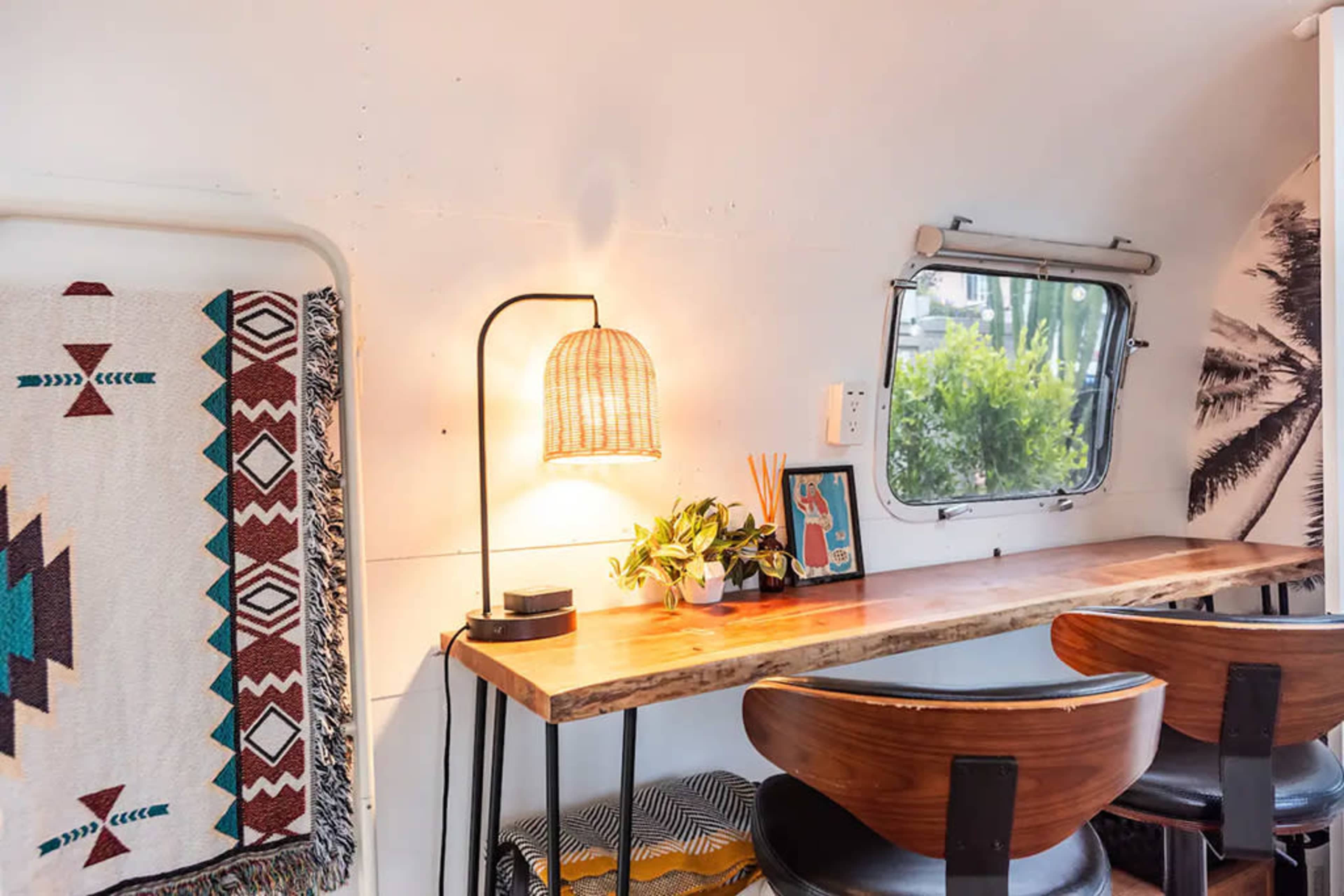 A cozy workspace is set up with a wooden desk, a lamp, and a small plant, surrounded by a blanket and wall decorations.