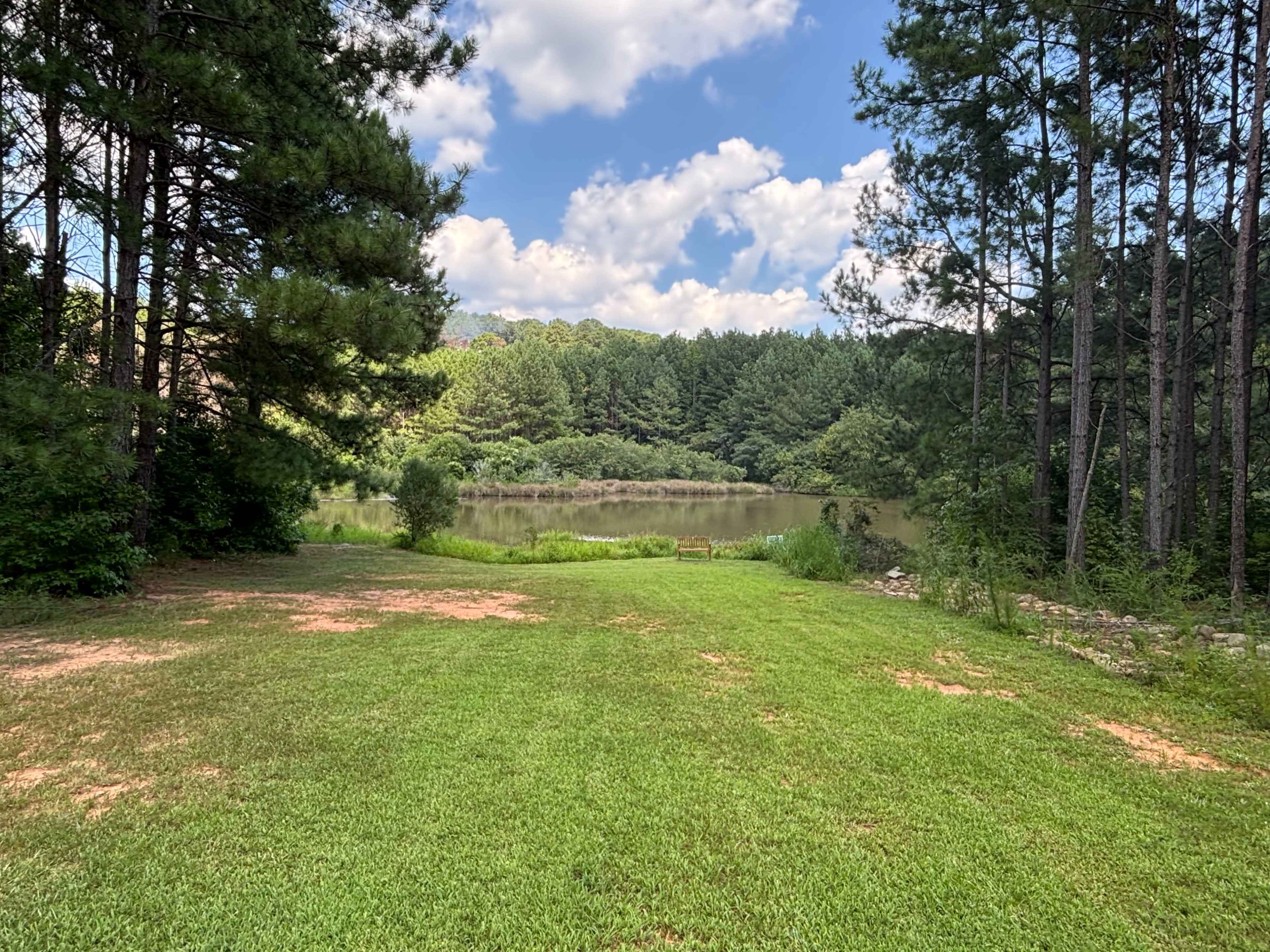 The image shows a grassy area bordered by trees, overlooking a calm body of water and a forested hillside under a partly cloudy sky.