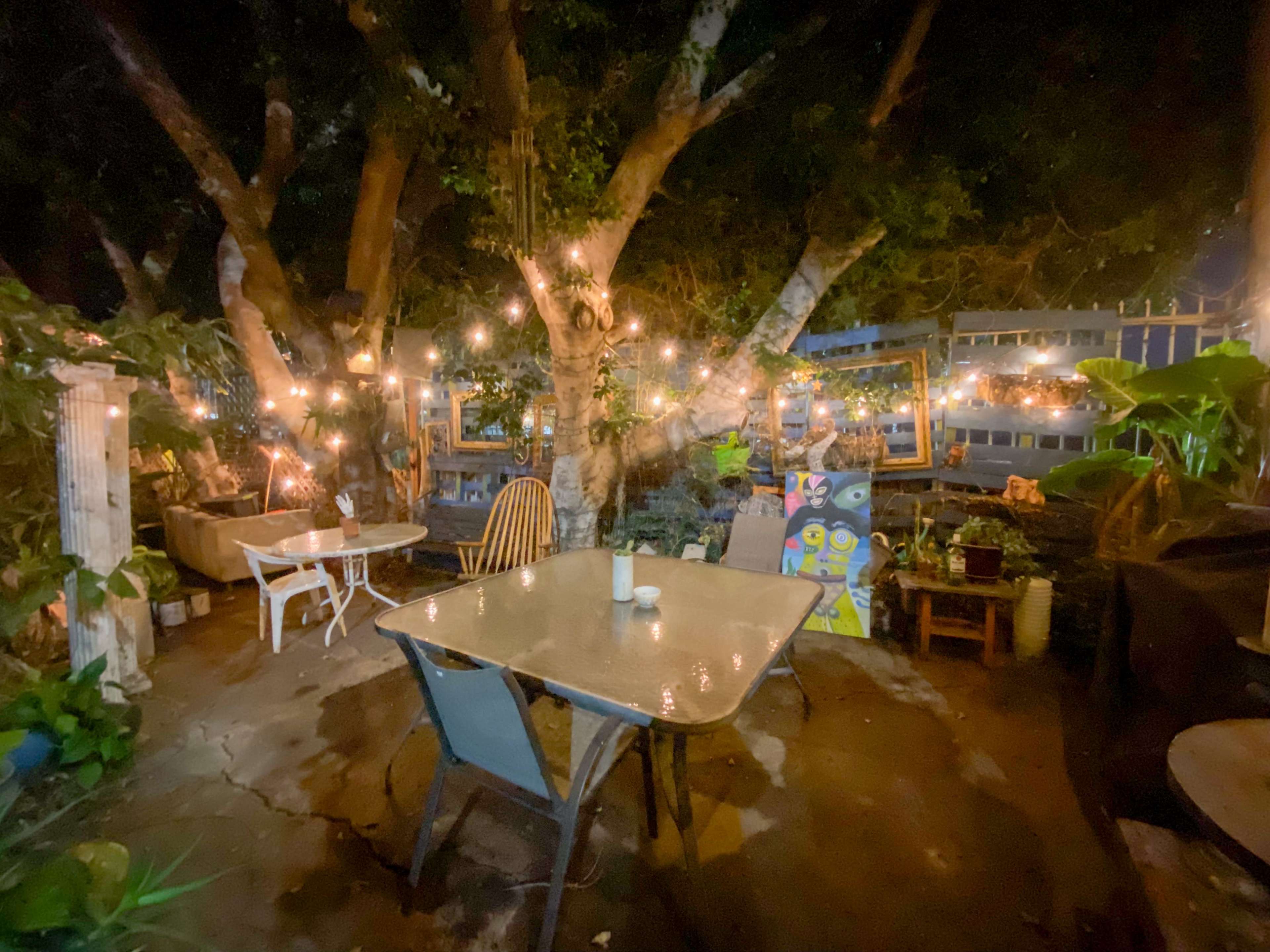 A garden patio at night is illuminated by string lights, featuring a table and chairs surrounded by trees and decorative artwork.
