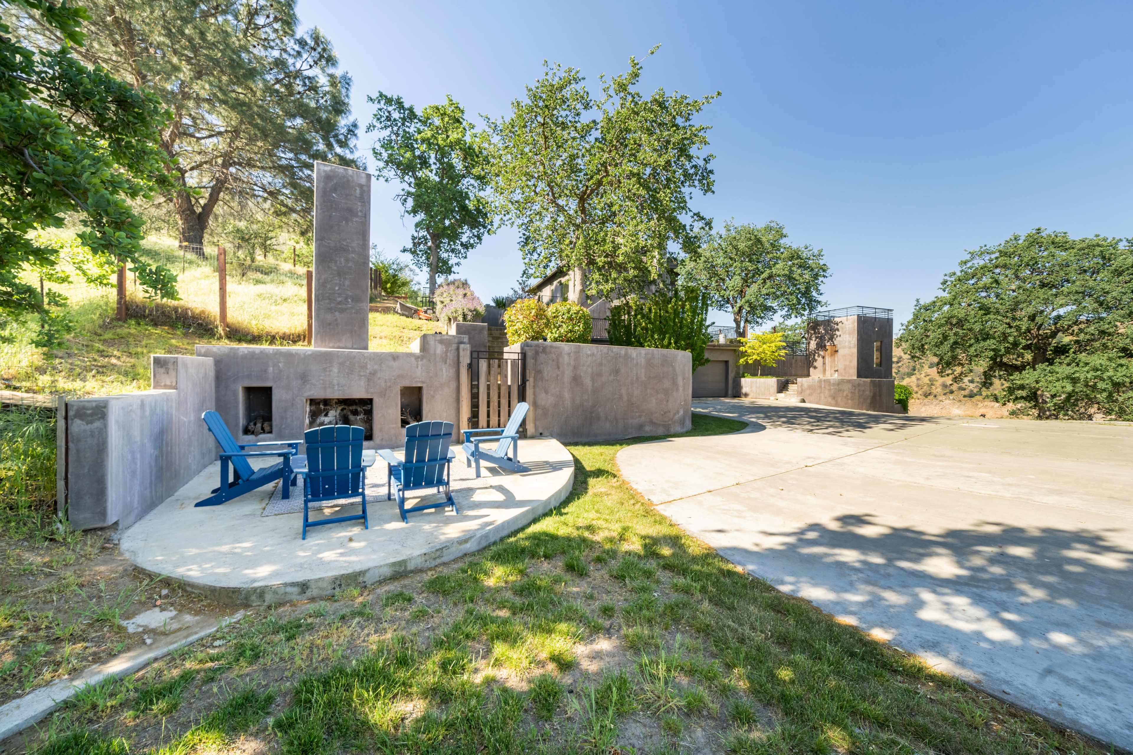 The image shows a modern outdoor space featuring a circular concrete patio with blue Adirondack chairs, surrounded by greenery and abstract architectural structures.
