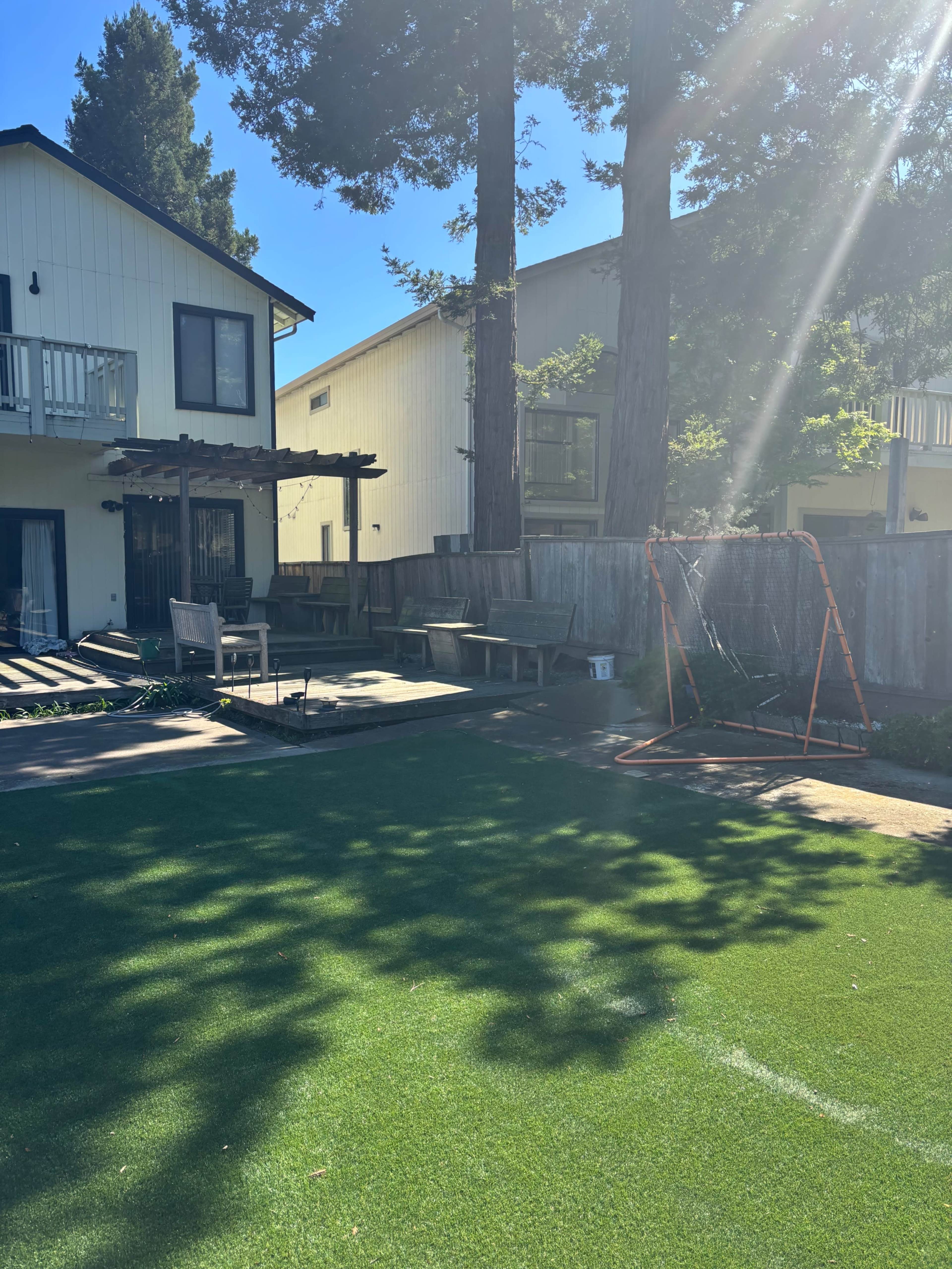 The image shows a backyard with artificial grass, a wooden deck, and a sports goal, surrounded by trees and residential buildings.