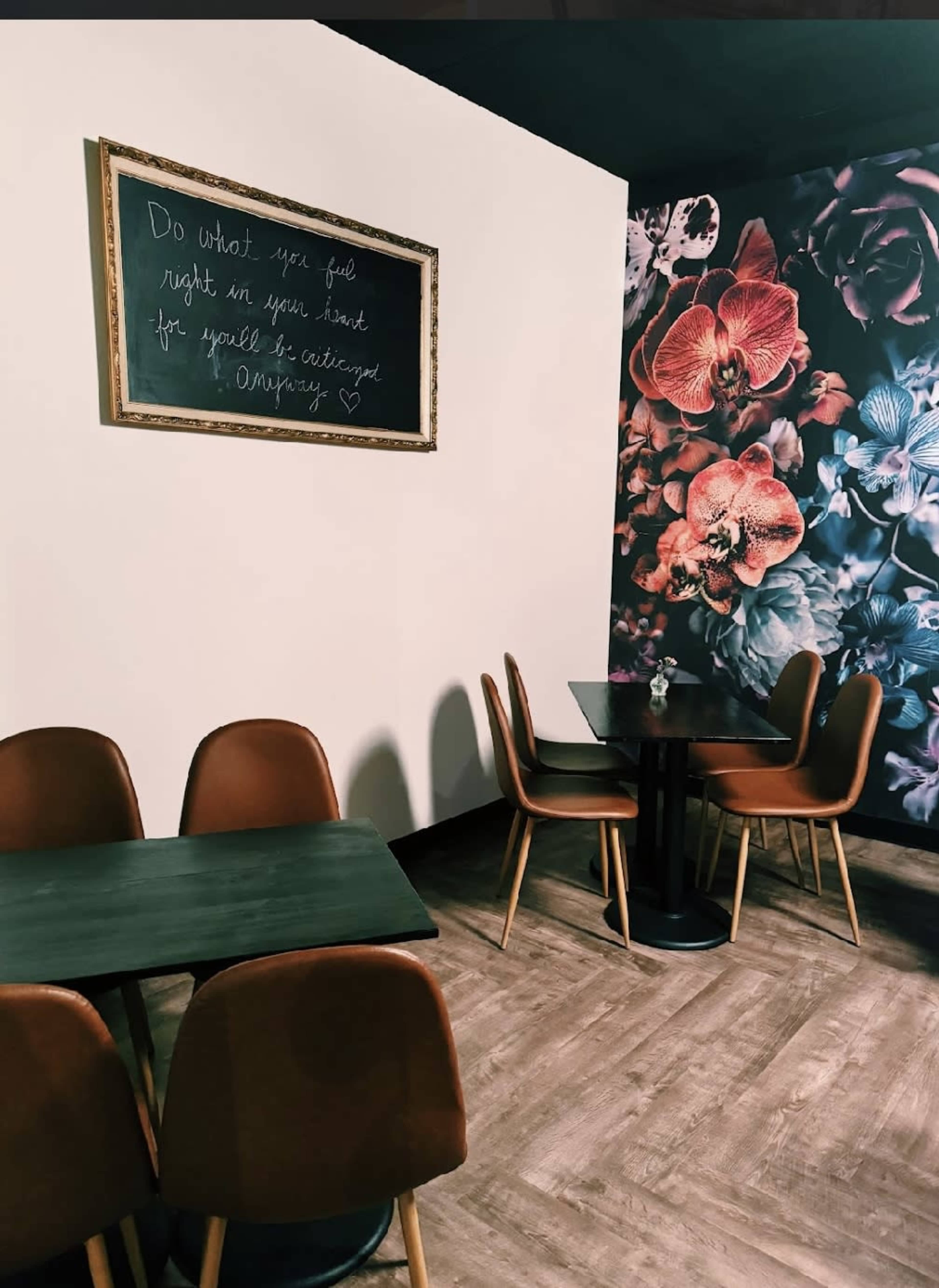 The image shows a cozy café interior featuring a floral-themed accent wall, wooden flooring, and stylish brown chairs around black tables.