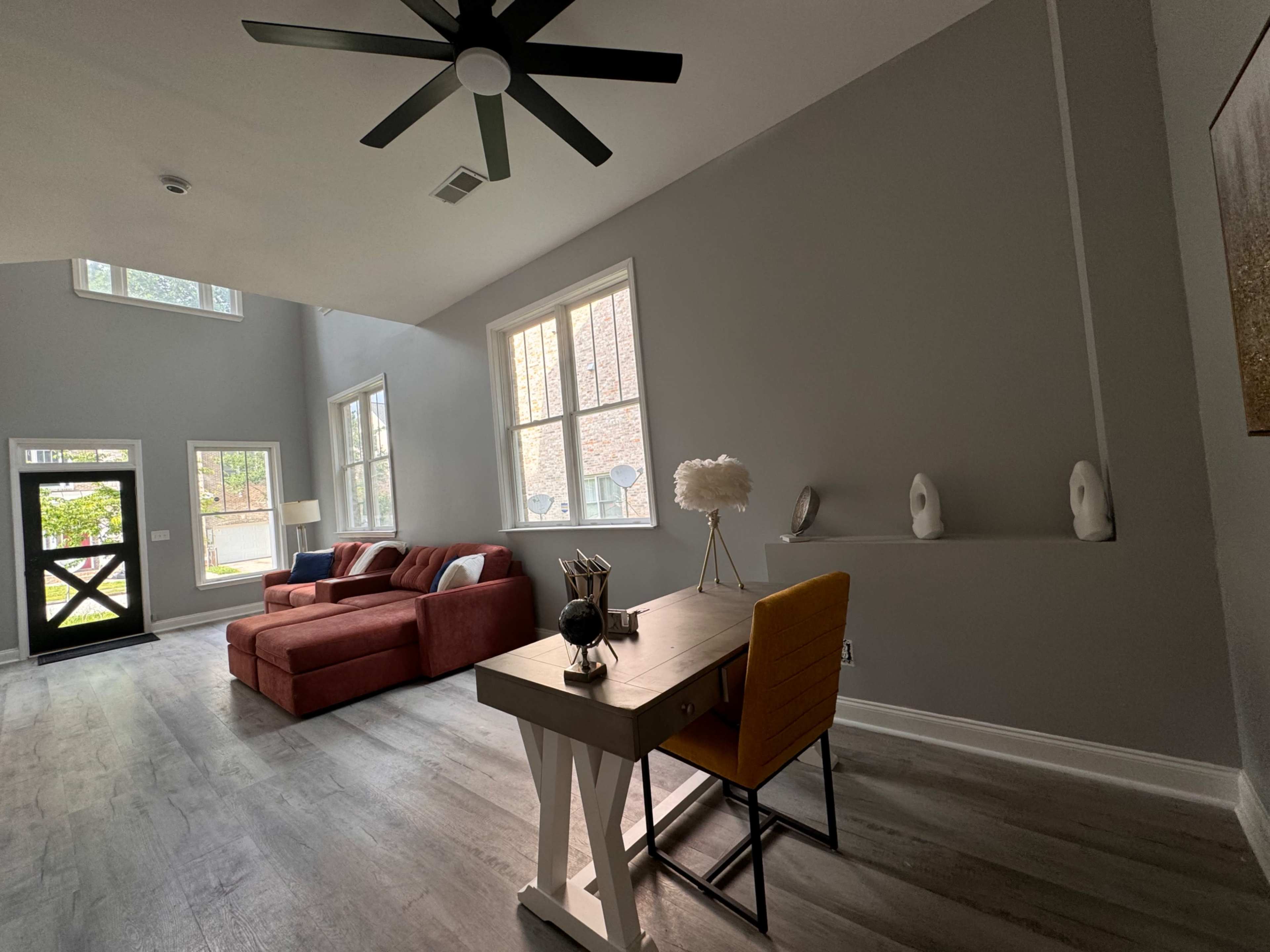 A spacious living area features a red sofa, a modern desk with a chair, and large windows allowing natural light to enter.
