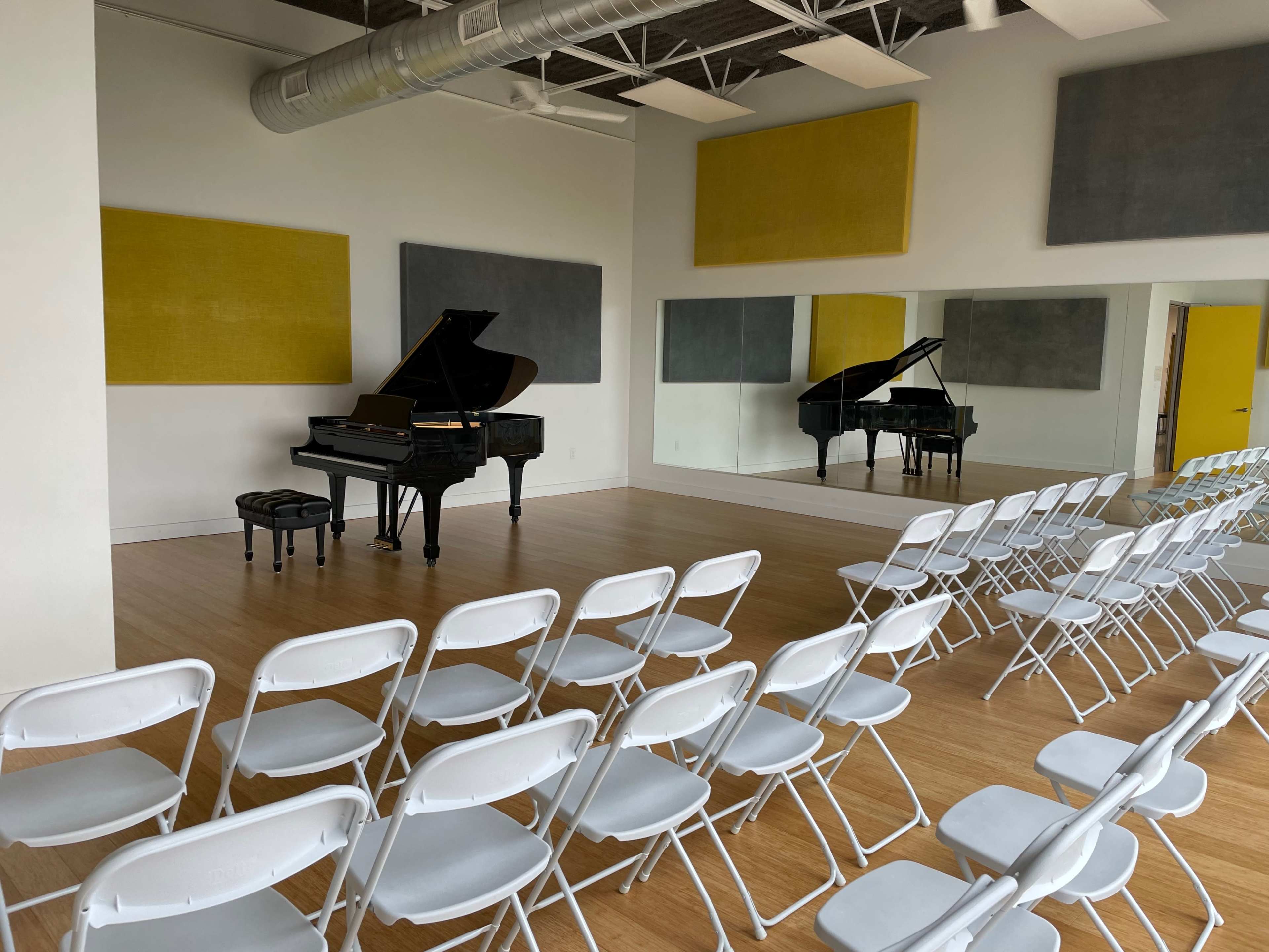 The image shows a music rehearsal space featuring two grand pianos, a small table with two chairs, and rows of white folding chairs arranged for an audience.