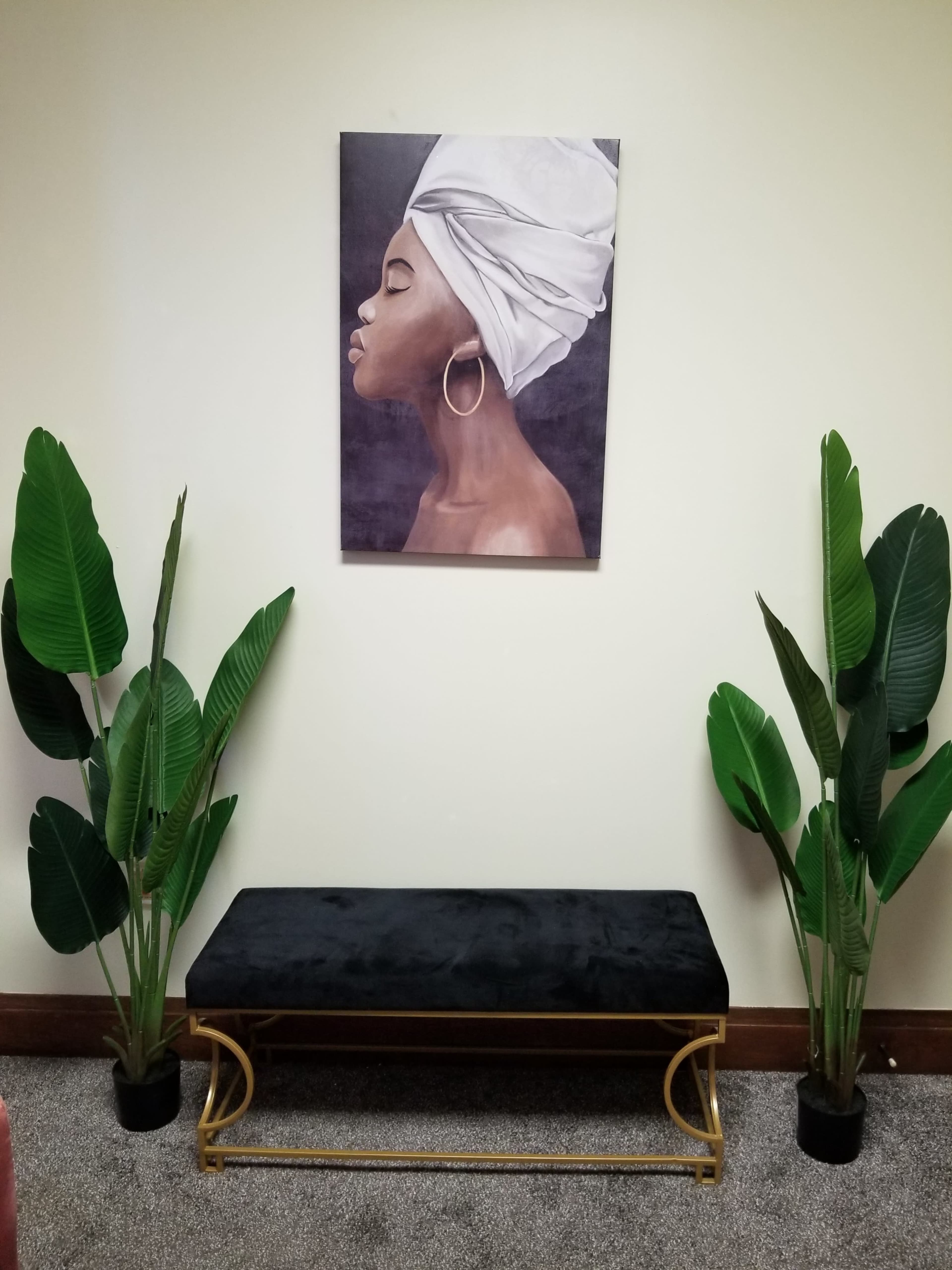 A black velvet bench is positioned between two tall green plants, with a portrait of a woman wearing a white head wrap hung on the wall above it.