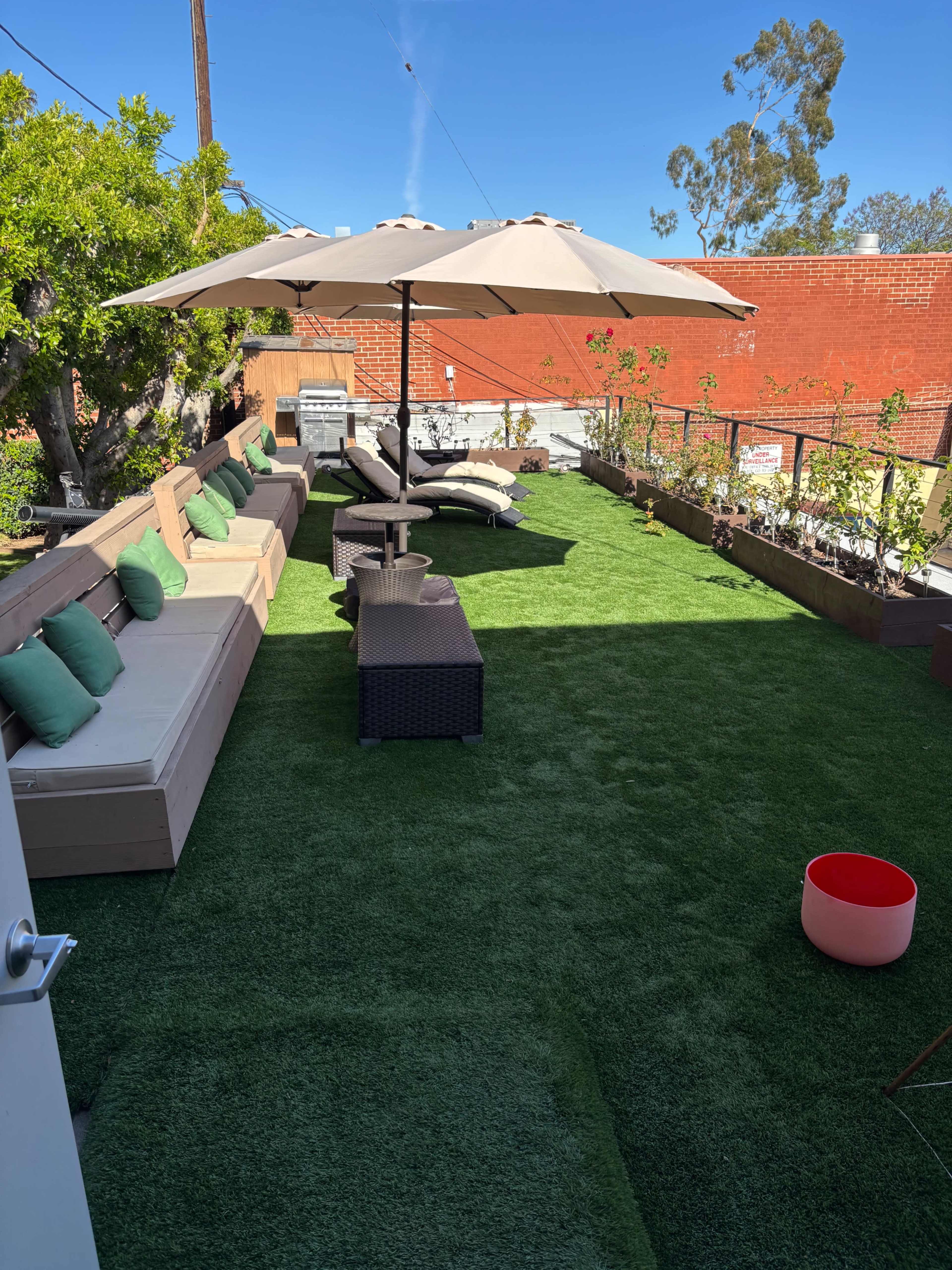 The image shows a rooftop patio featuring artificial grass, several lounge chairs with cushions, an umbrella for shade, and potted plants along the perimeter.