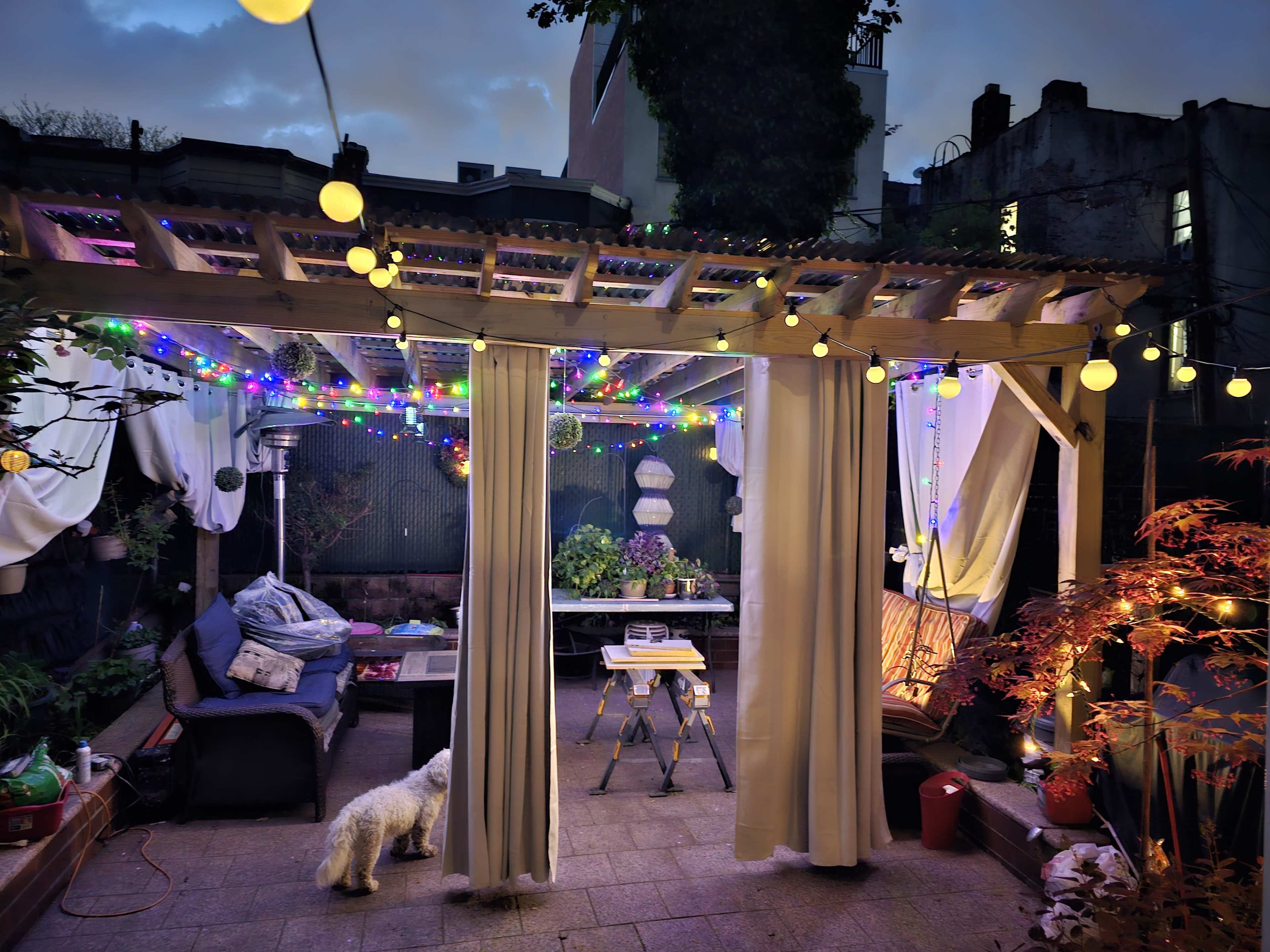 The image shows a backyard patio with a wooden pergola adorned with colorful string lights and curtains, featuring a table and seating area along with a small dog.