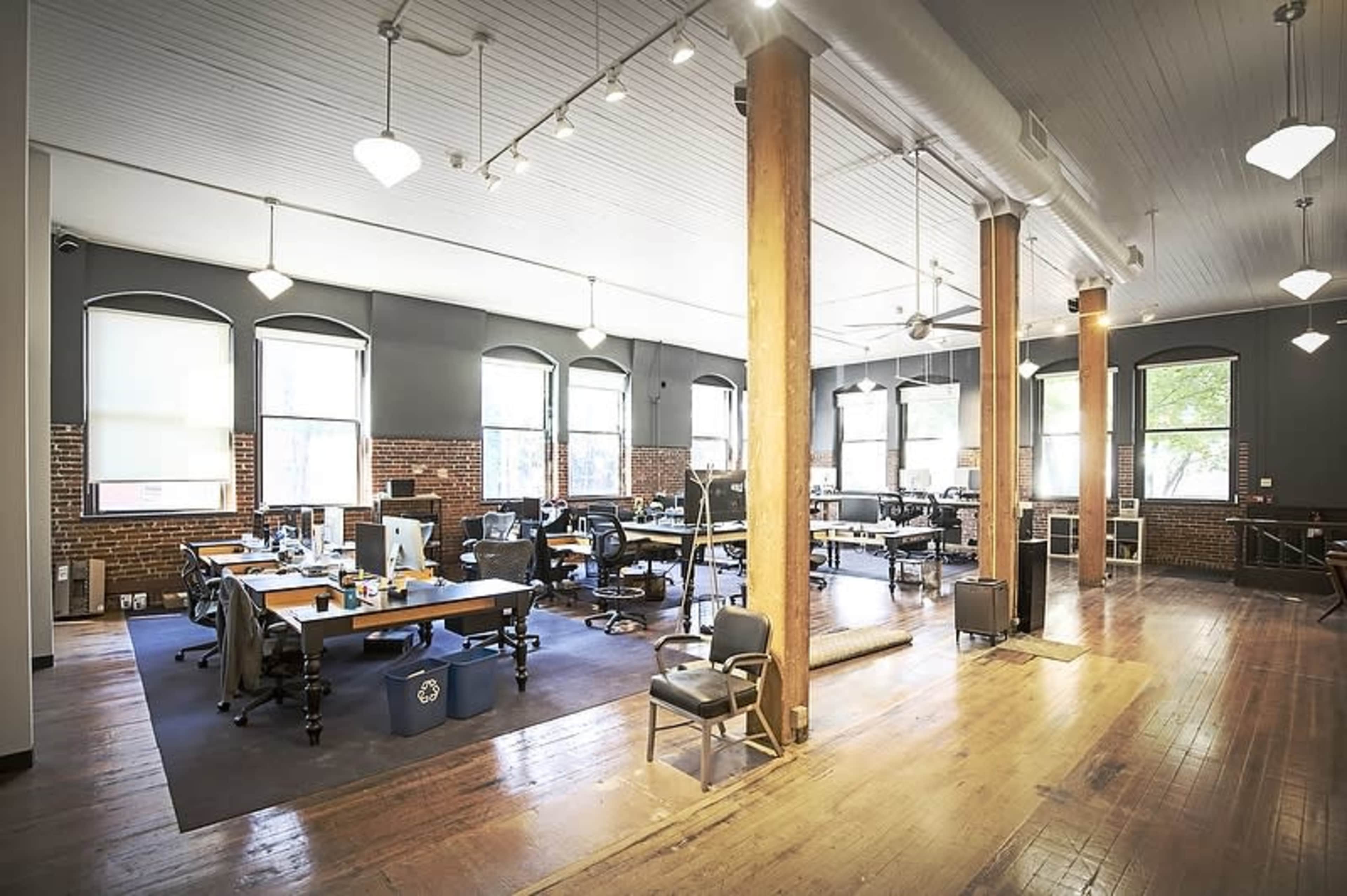A spacious office with wooden floors, large windows, and rows of desks equipped with computers.