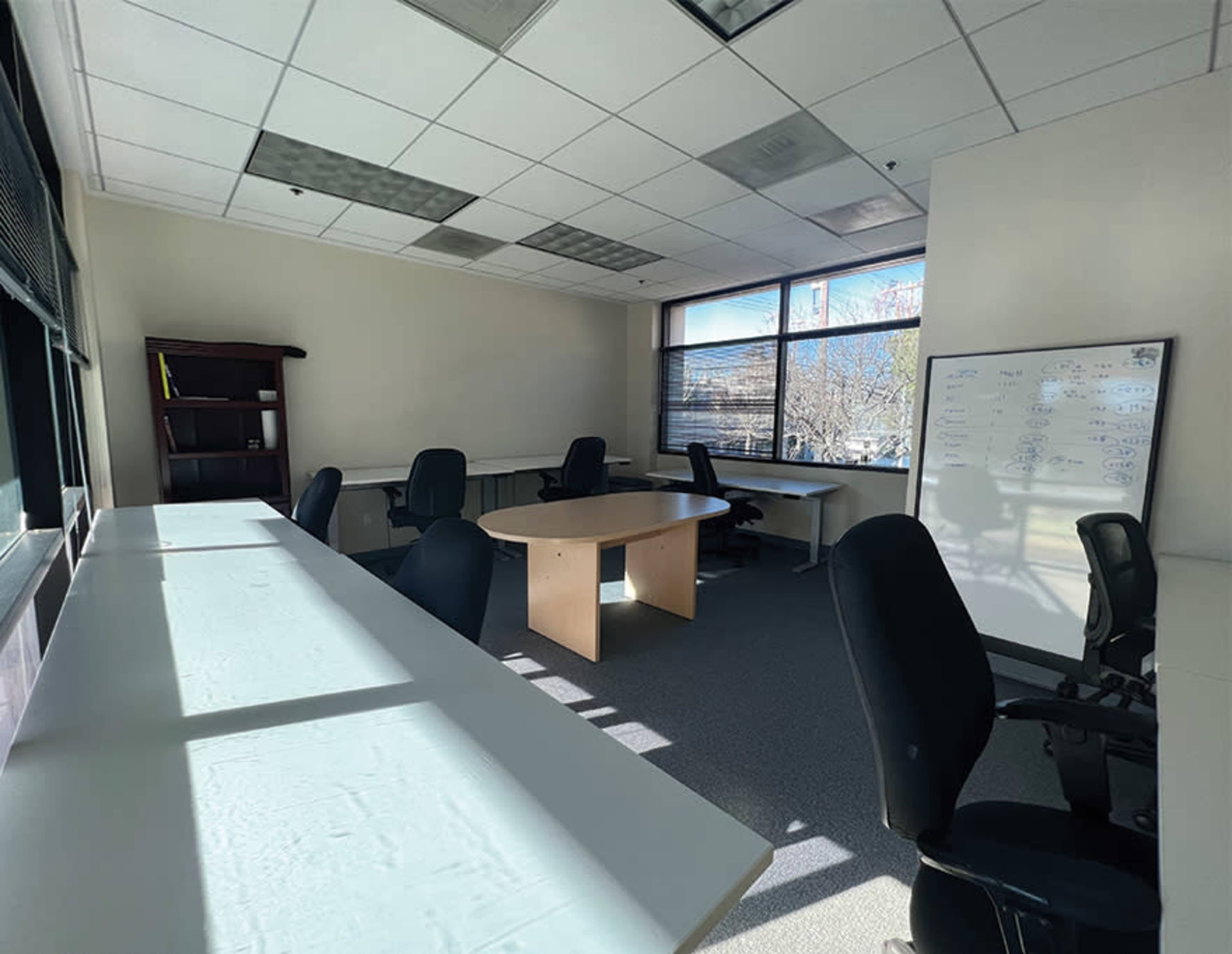 A spacious office with several desks, chairs, and a whiteboard, illuminated by natural light coming through a large window.