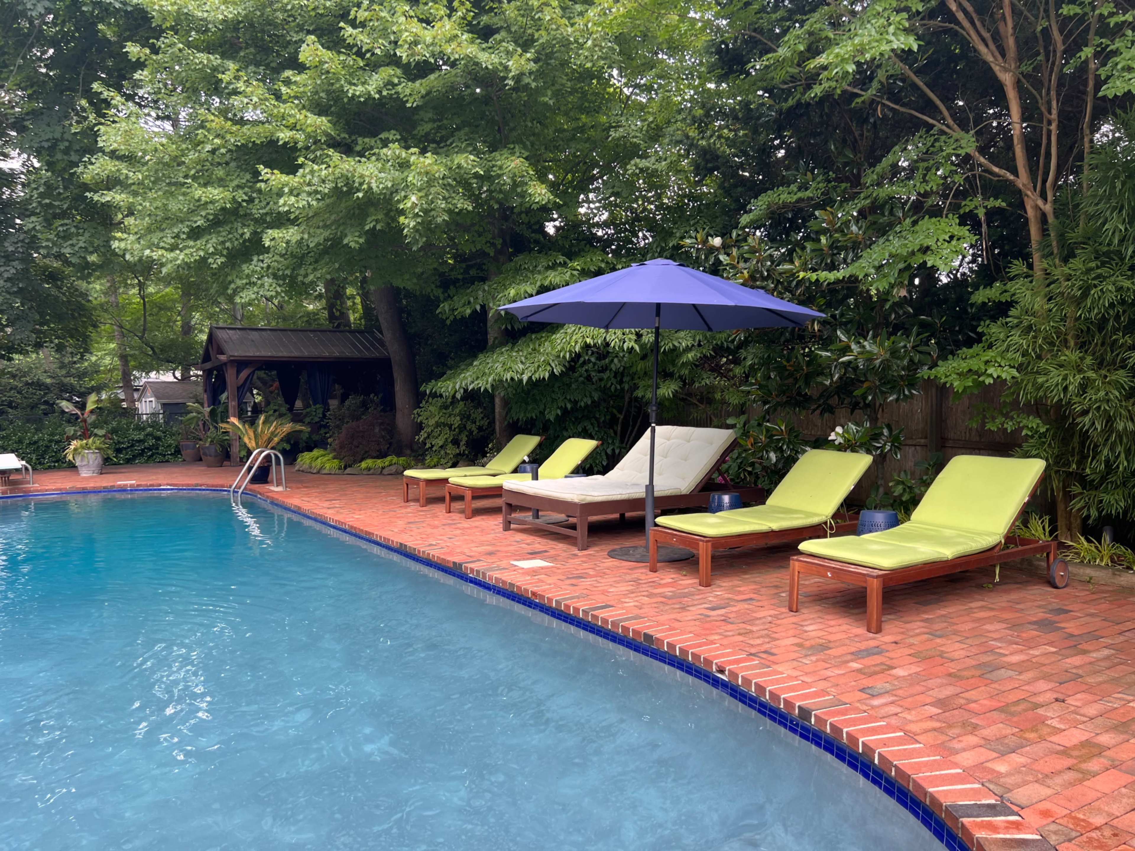 The scene shows a swimming pool surrounded by brick decking, with several green lounge chairs and a large umbrella positioned nearby.
