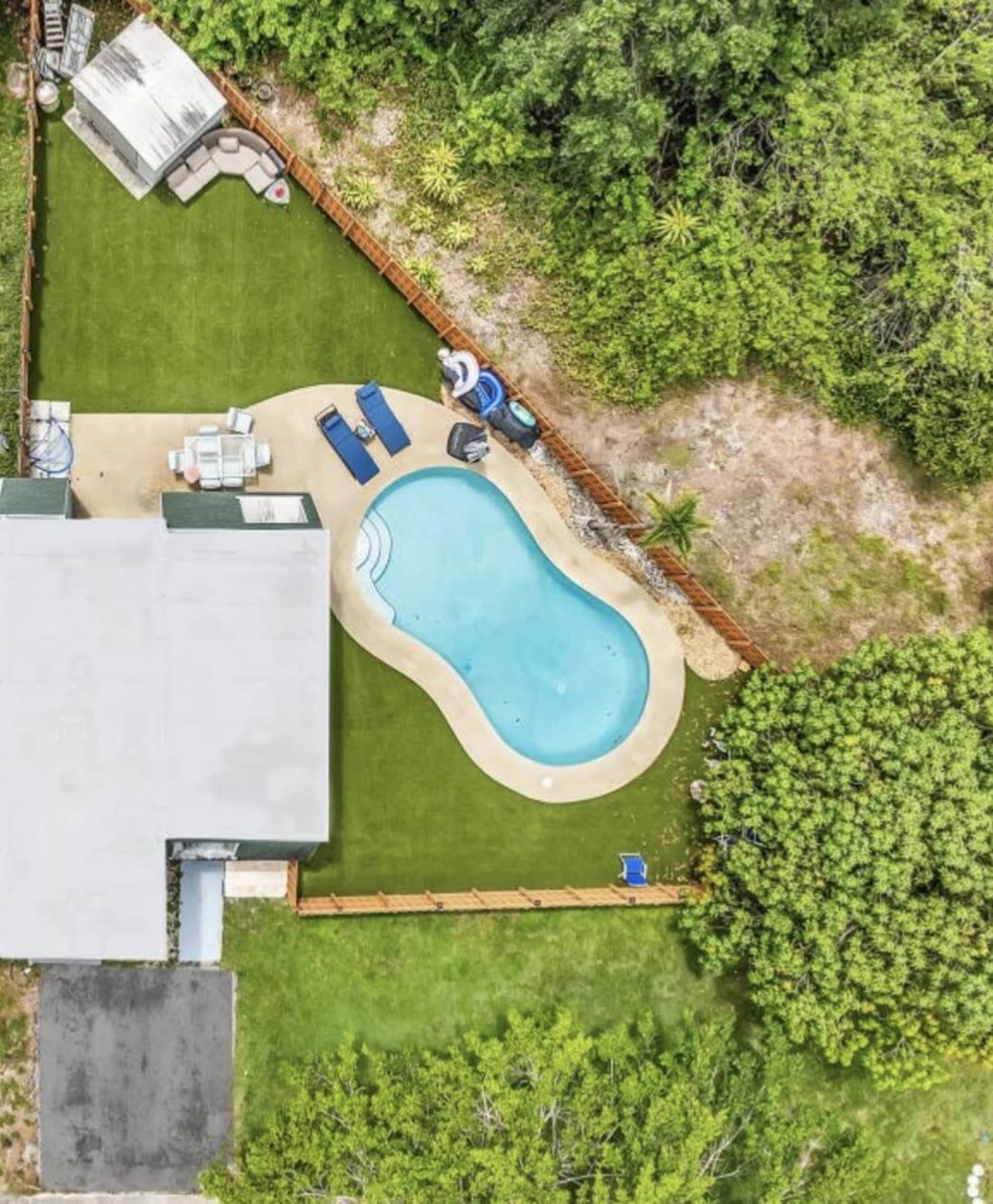 The image shows a backyard with a swimming pool, surrounded by green grass and a wooden fence, adjacent to a house and some wooded area.