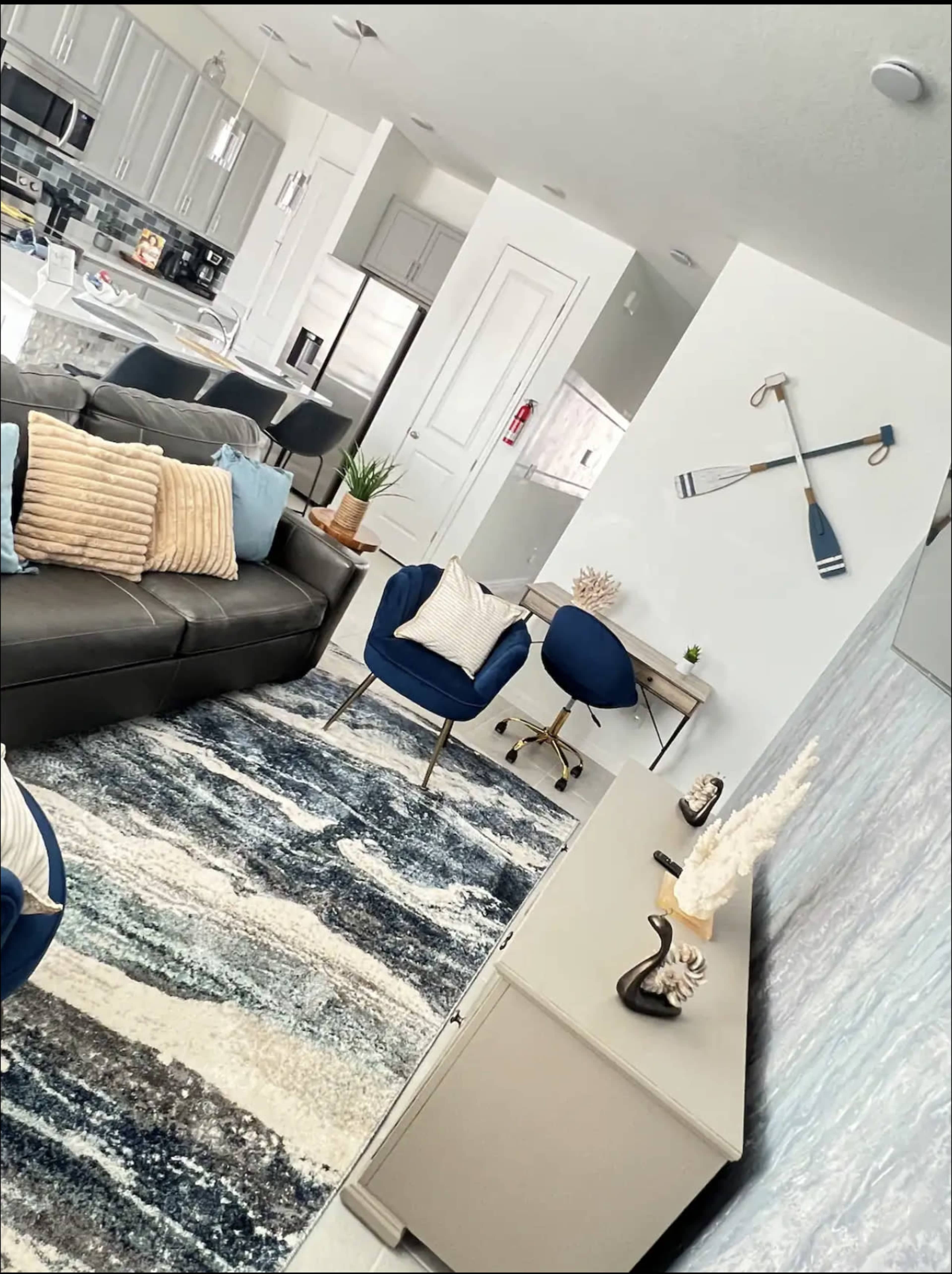 The image shows a modern living room with a gray sofa, blue accent chairs, and a decorative wall featuring oars, along with a light-colored area rug.