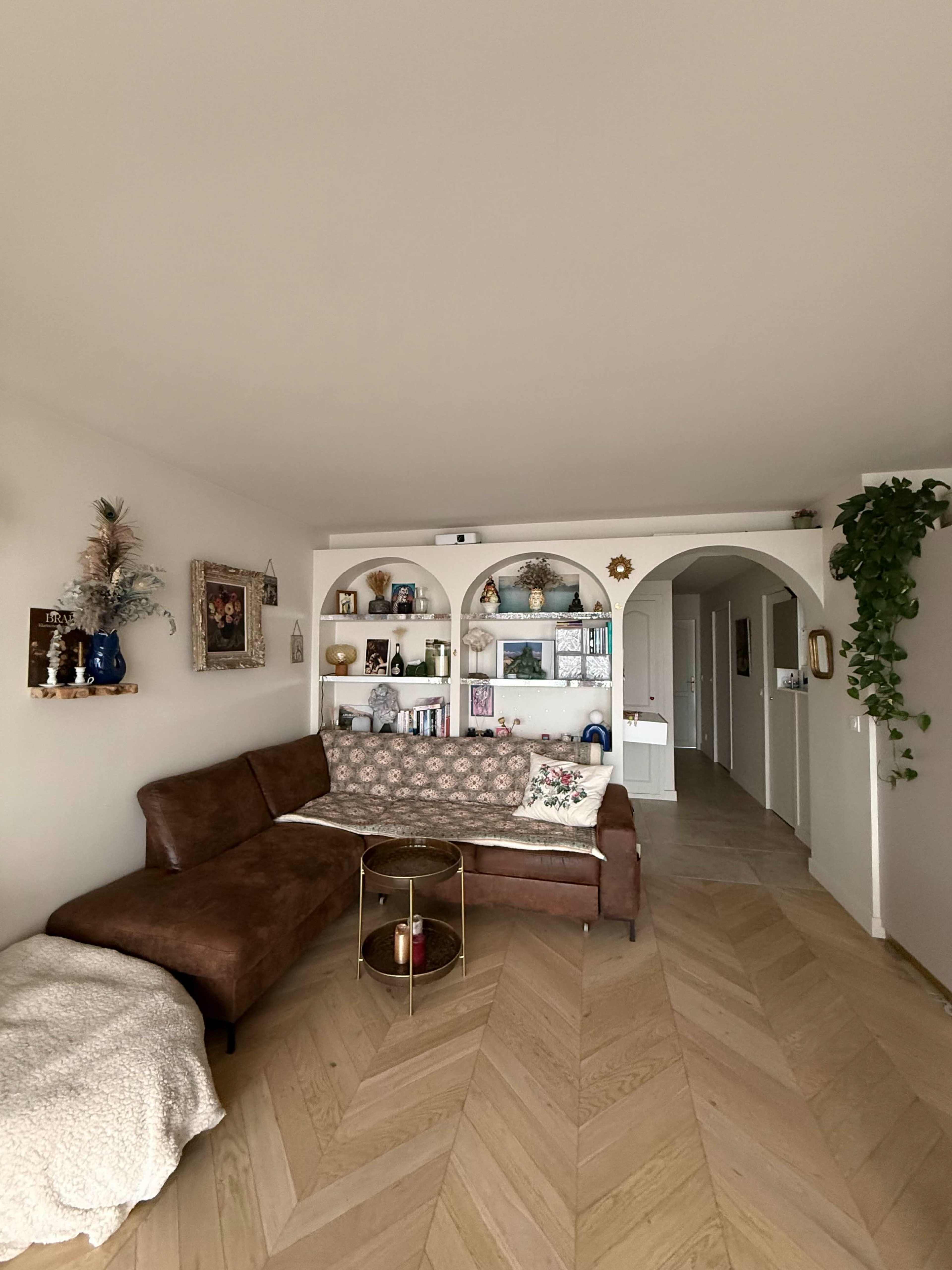 The image shows a modern living room with a brown sectional sofa, a decorative shelving unit, and a light wood herringbone floor.