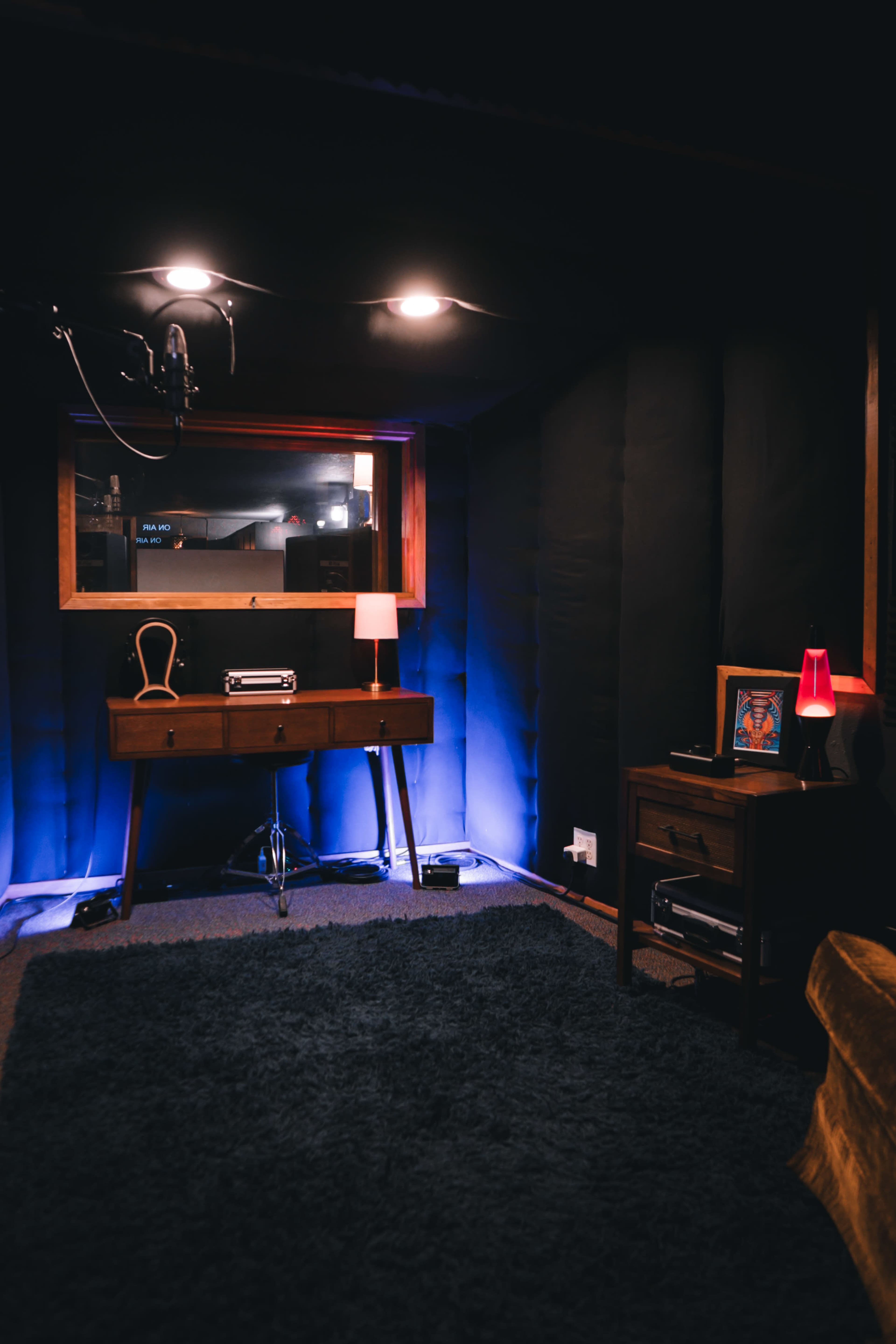 The image shows a dimly lit recording studio with acoustic panels, a microphone overhead, a small table with a lamp, and a window looking into a control room.