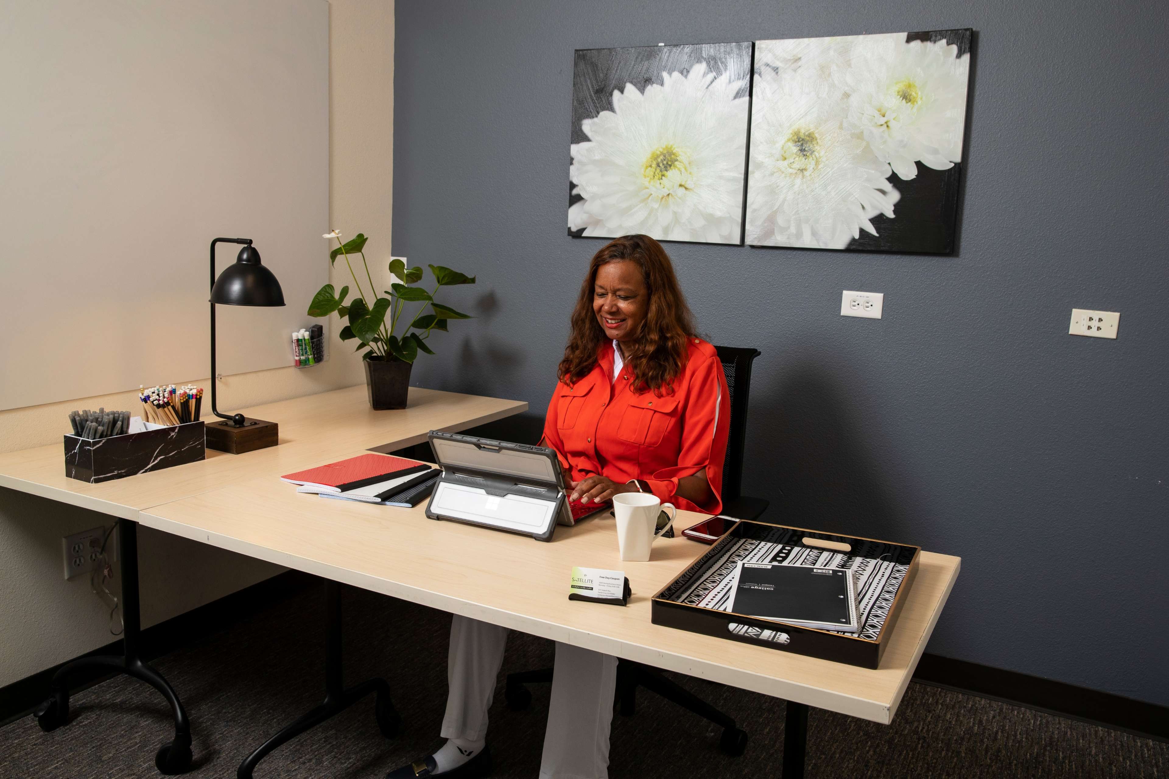 A woman in an orange blouse sits at a desk working on a tablet in an office with a flower-themed wall decoration.