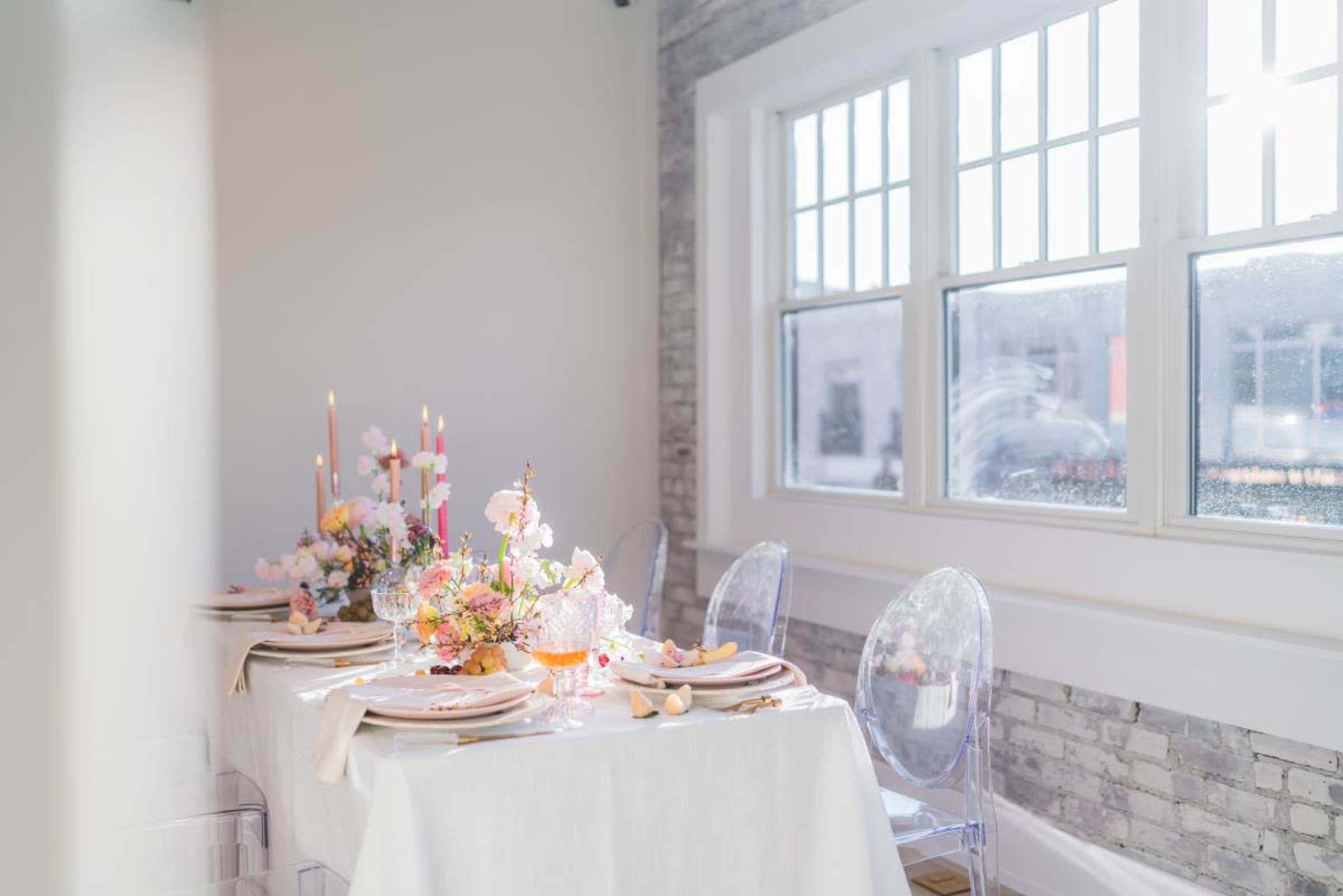 A beautifully set dining table with floral arrangements, candles, and elegant tableware is positioned by a large window.