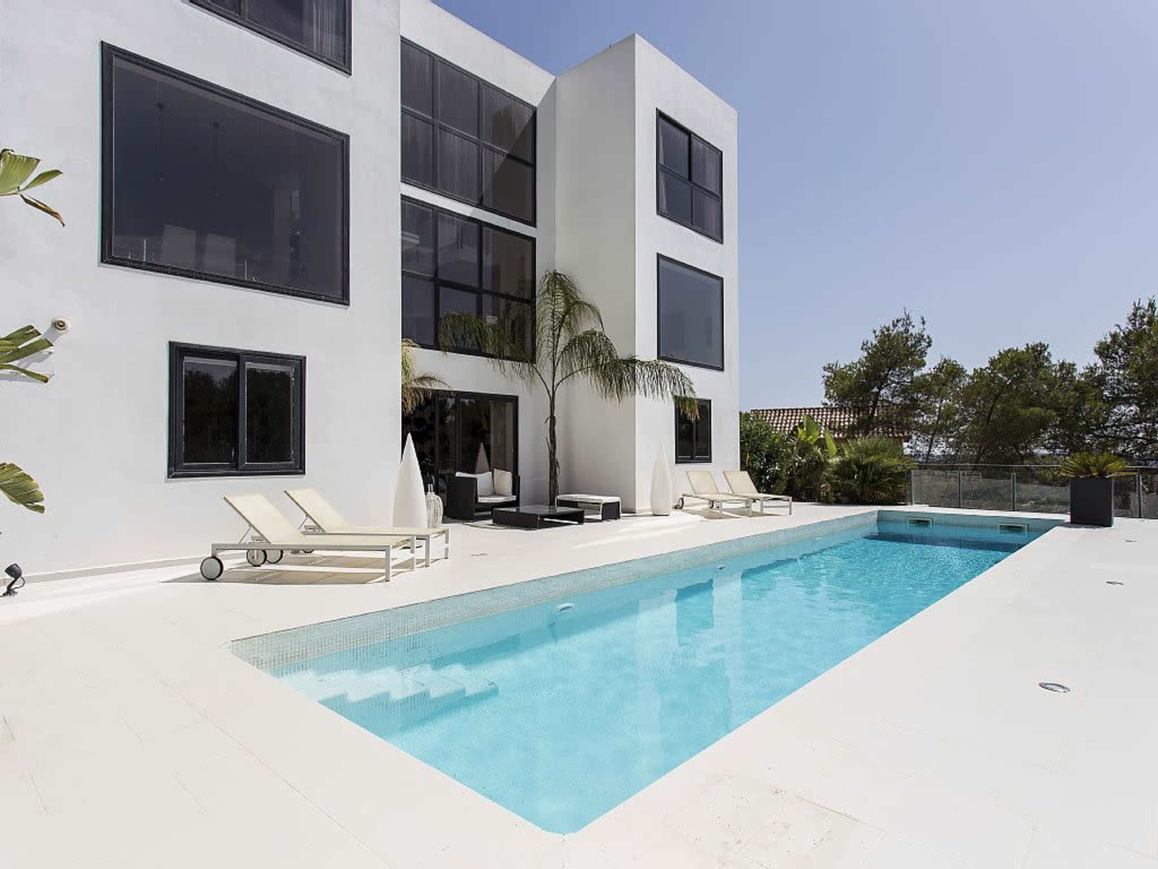 A modern white villa with large windows overlooks a rectangular swimming pool surrounded by lounge chairs and tropical plants.