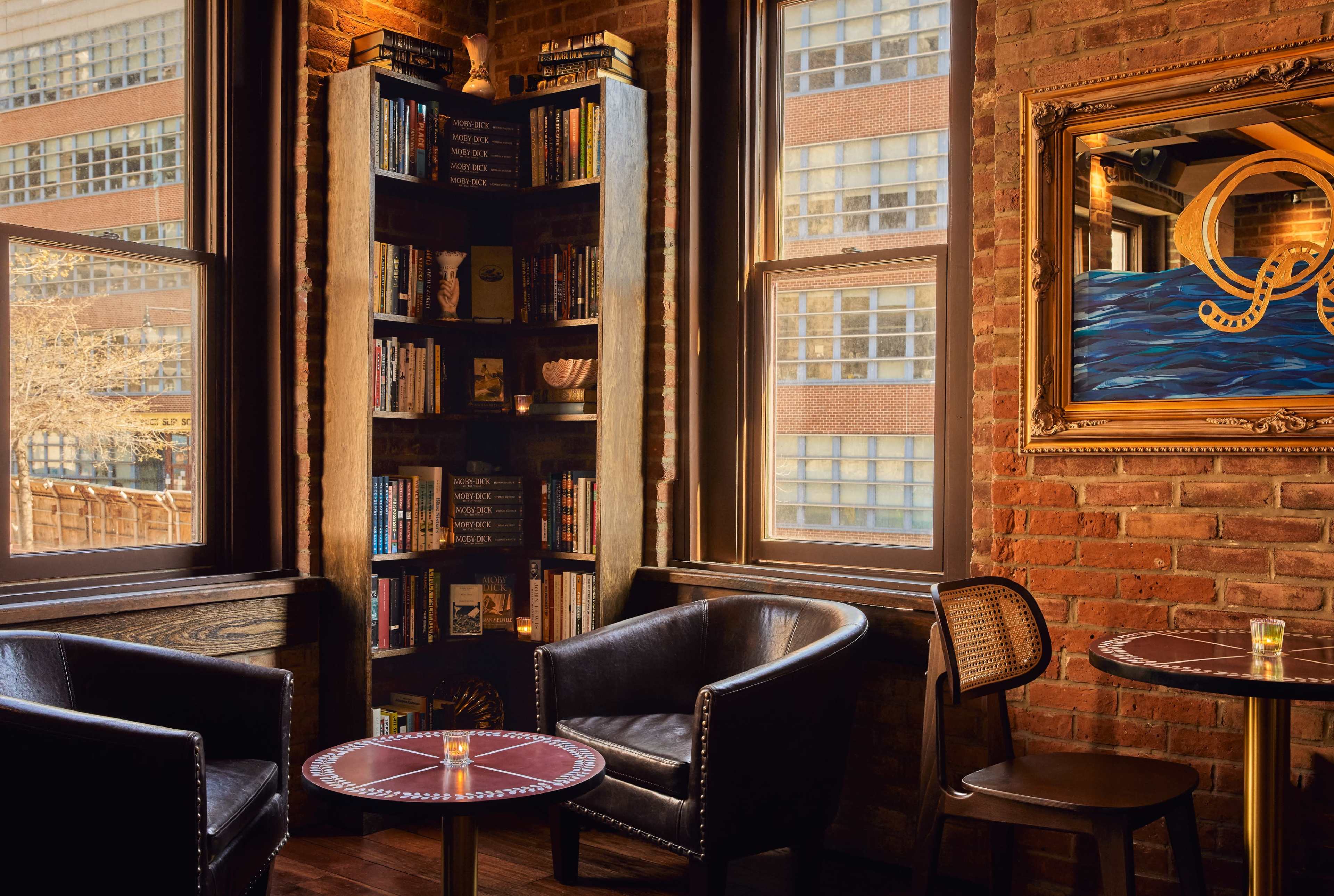 A cozy corner of a cafe features two black leather chairs and a small table next to a bookshelf filled with books and a large window offering a view of the outside.