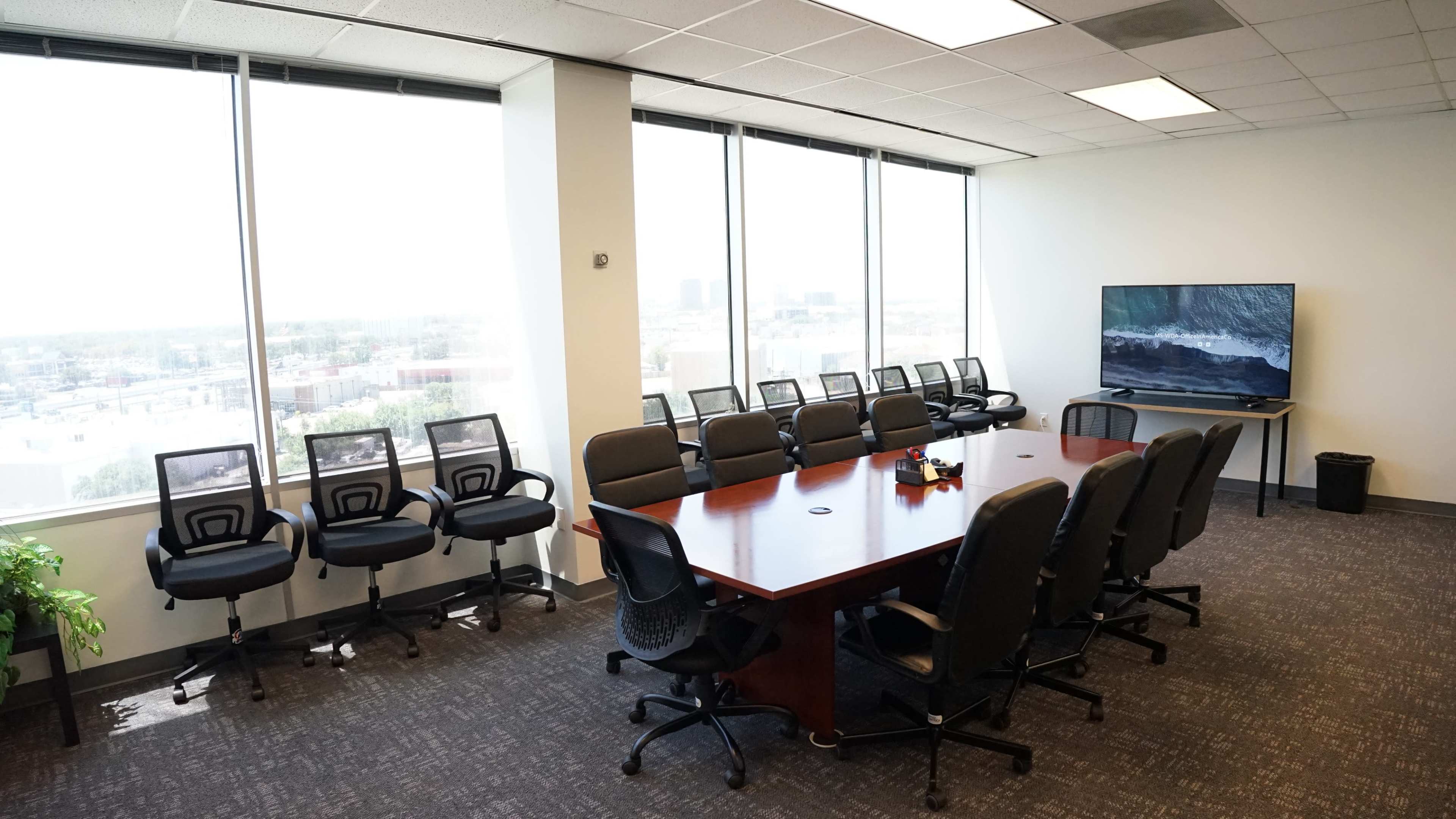 A conference room features a large wooden table surrounded by black office chairs, with floor-to-ceiling windows overlooking the city and a television mounted on the wall.