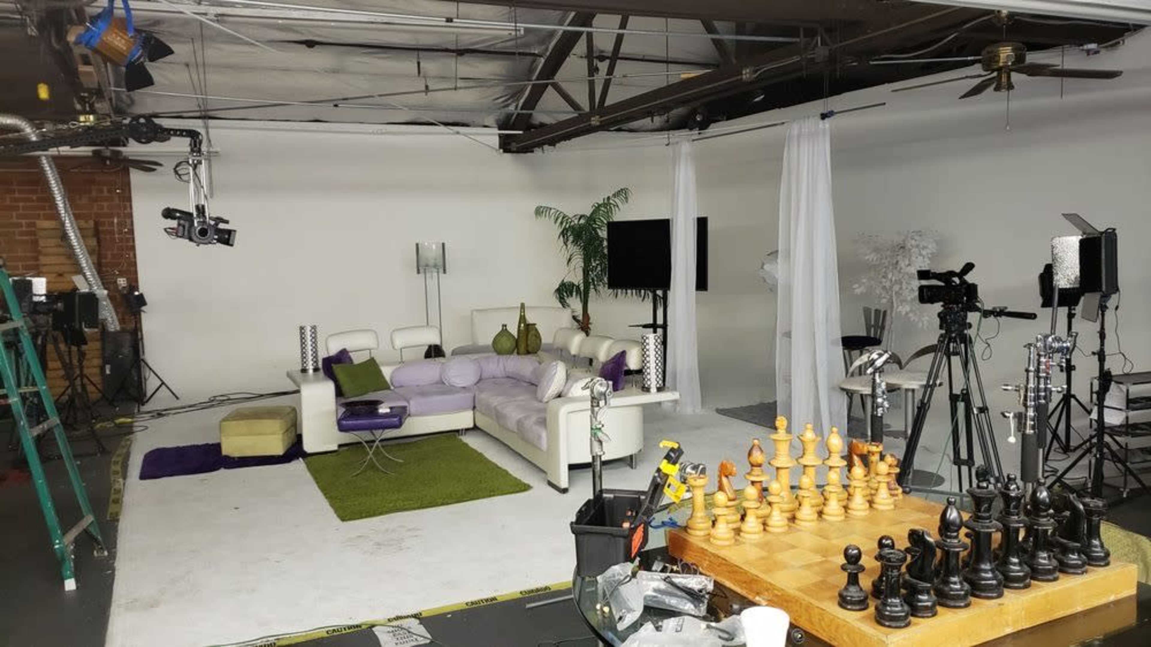 A studio space featuring a white couch with purple accents, a chess set in the foreground, and various cameras and lighting equipment arranged around the room.