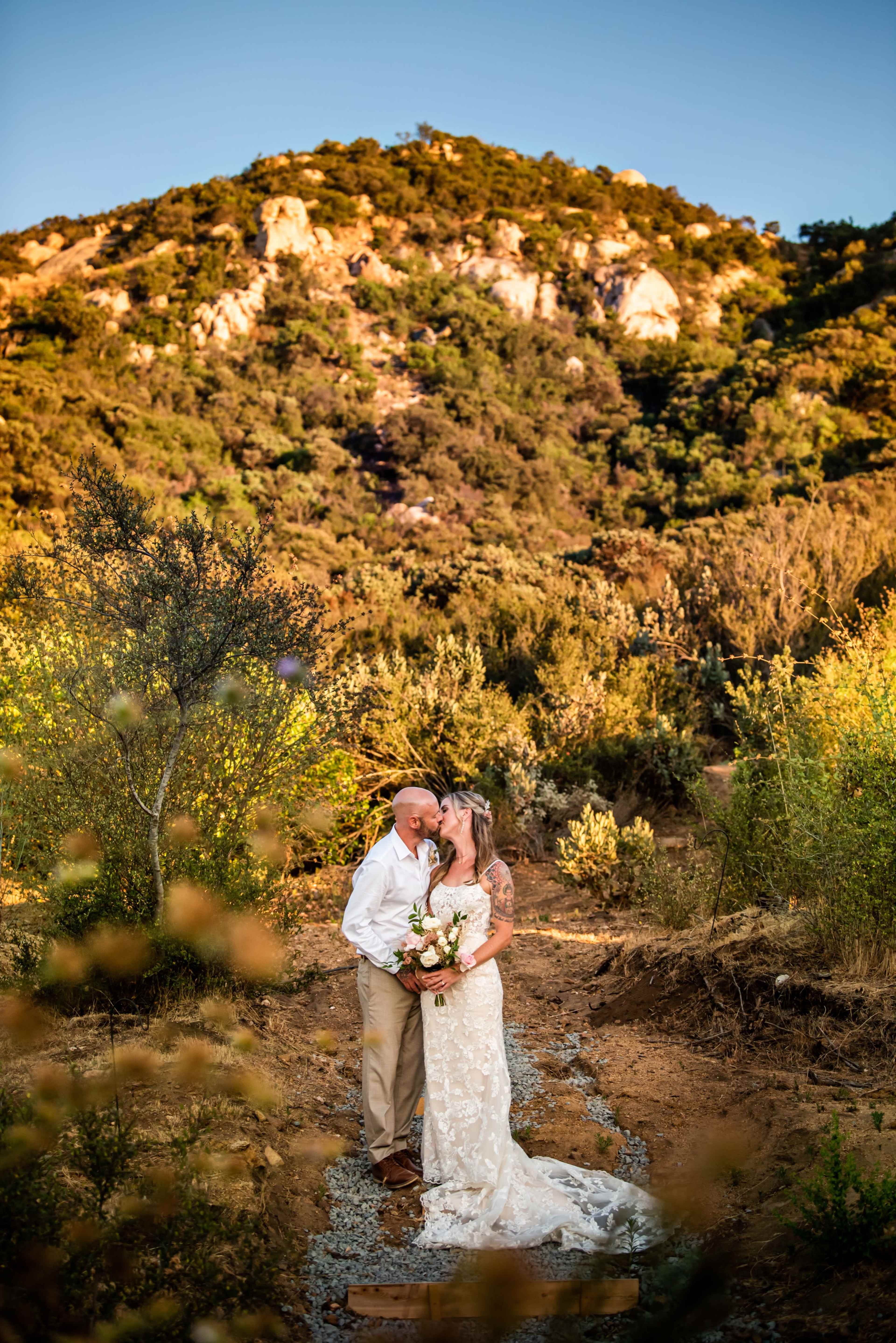 A couple in wedding attire embraces on a gravel path surrounded by greenery and a mountainous backdrop.