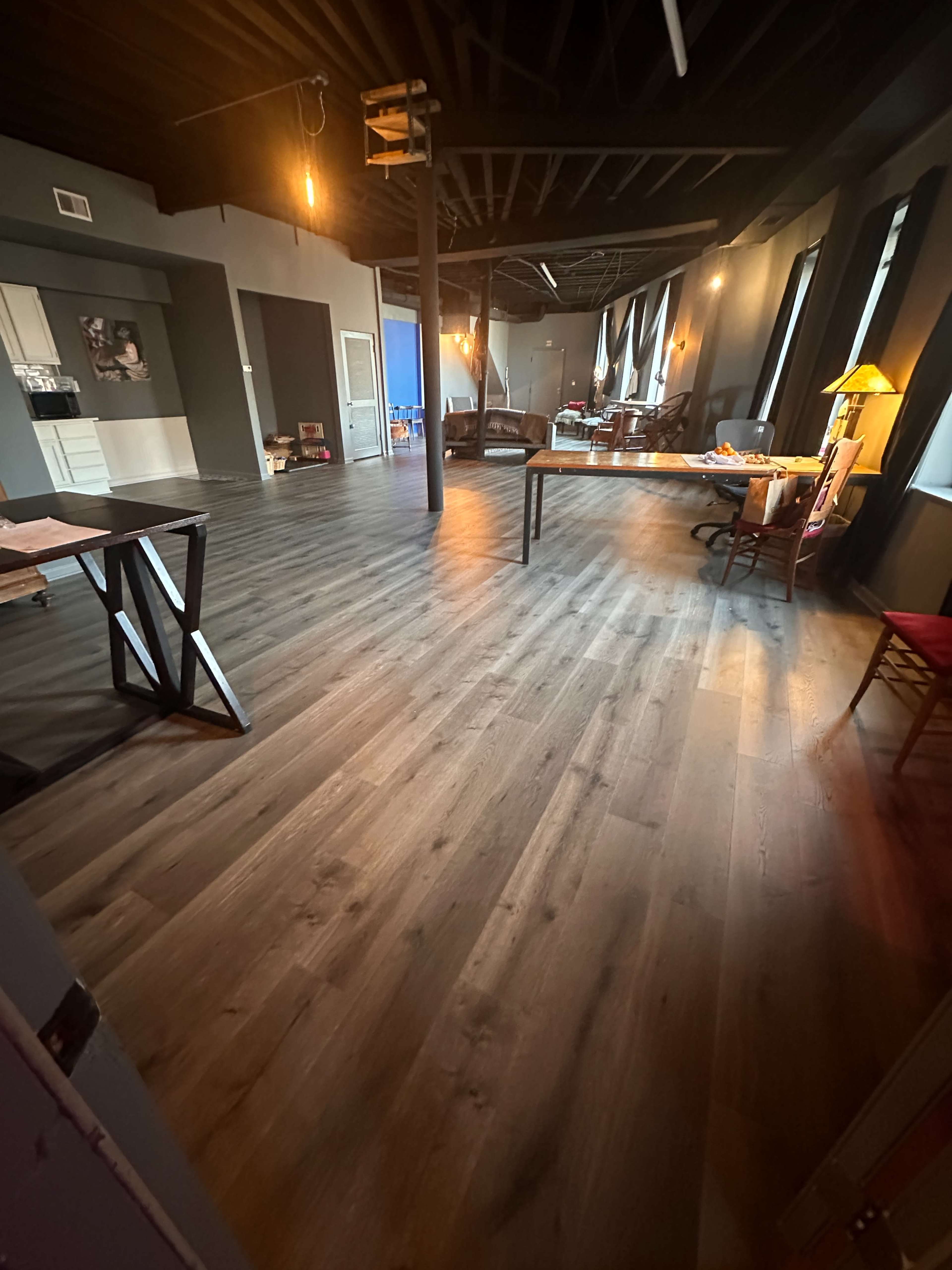 The image shows a spacious, modern office interior with wooden flooring, beams on the ceiling, and various furniture pieces scattered throughout.