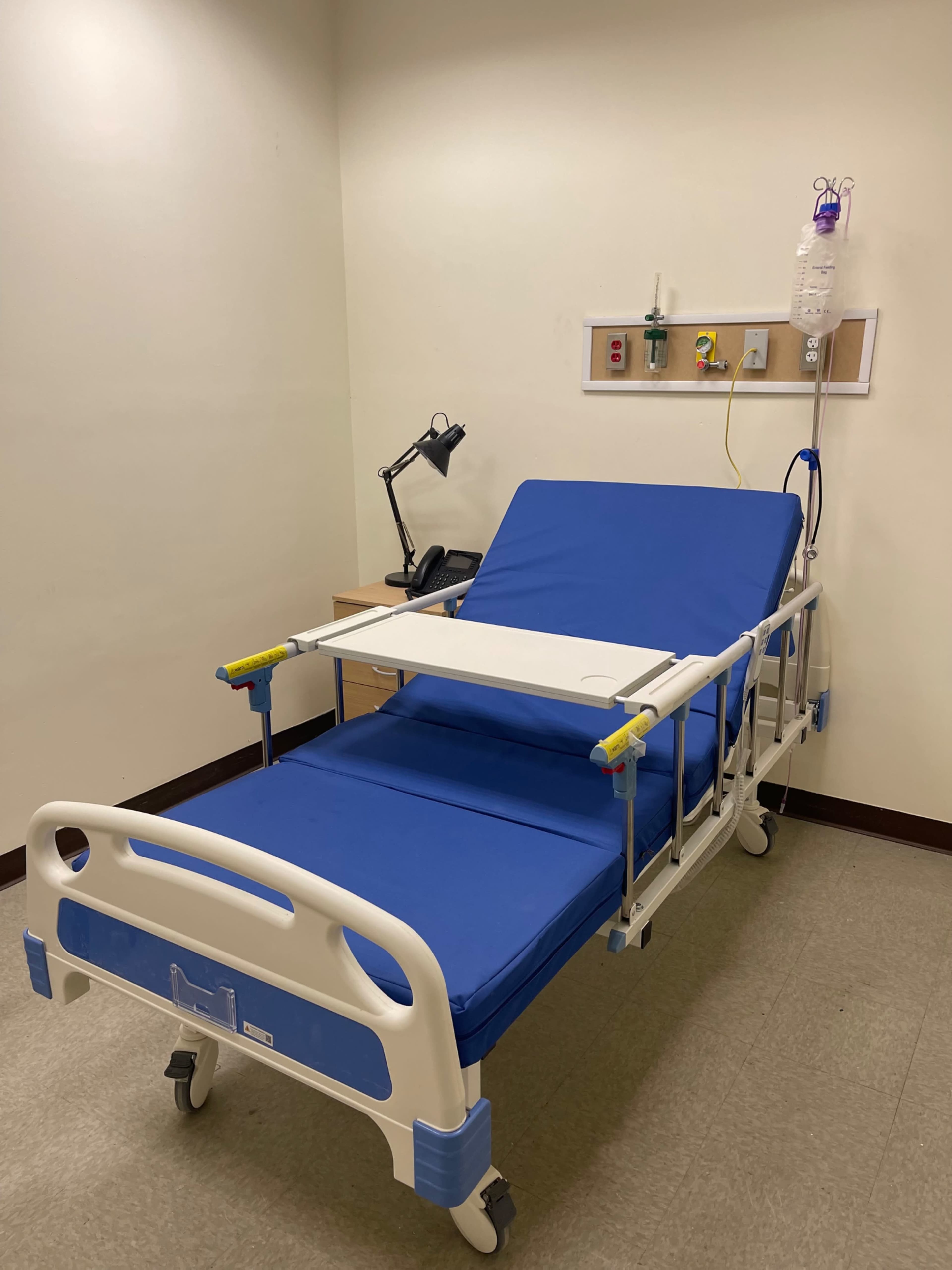 The image shows a hospital room with a blue hospital bed, a bedside table, a lamp, and an IV stand with a bag attached.