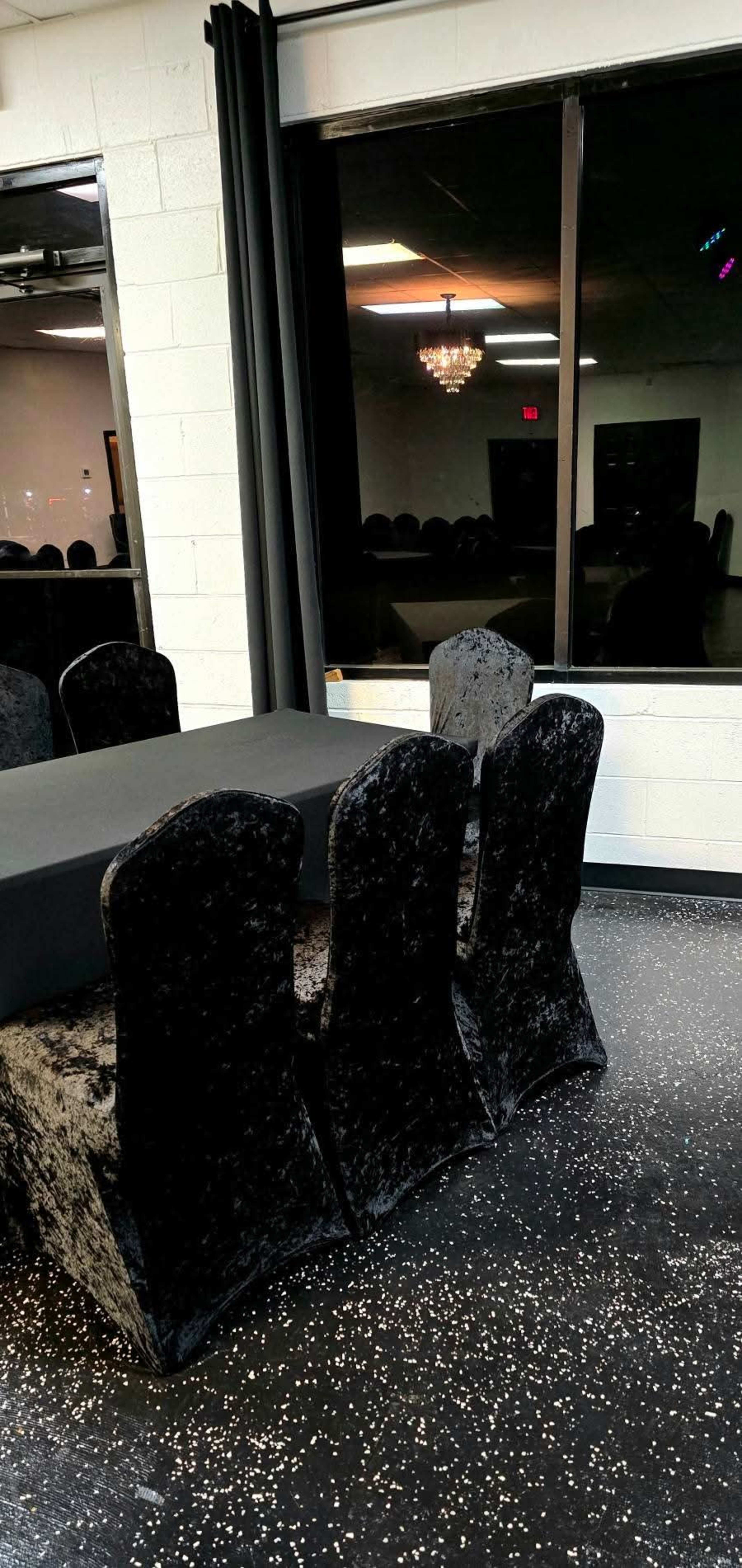 A black table with velvet chairs is set against a window reflecting a chandelier and the dimly lit interior of a room.