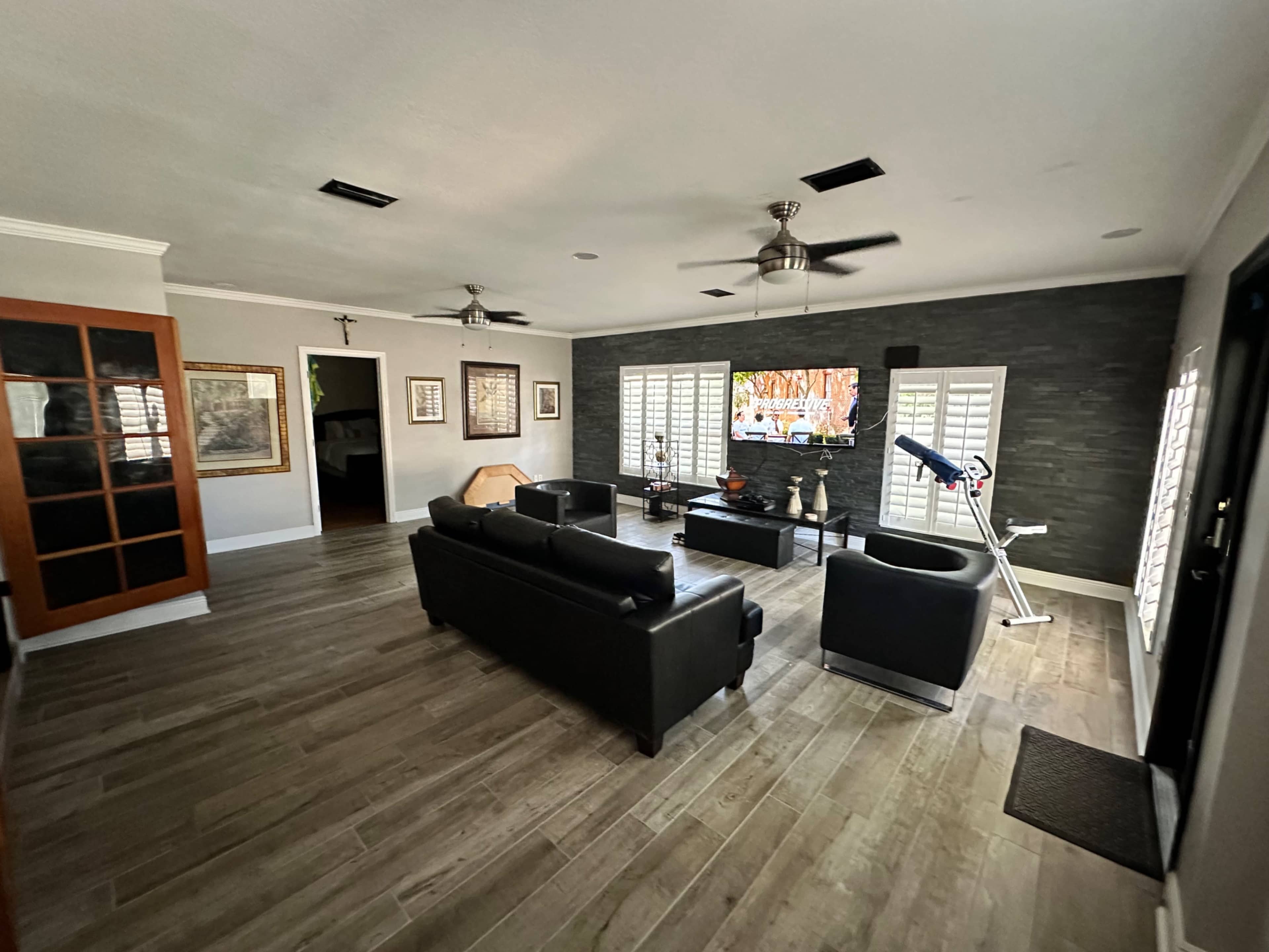 A spacious living room with dark leather furniture, a large flat-screen TV mounted on a stone wall, and exercise equipment in the corner.