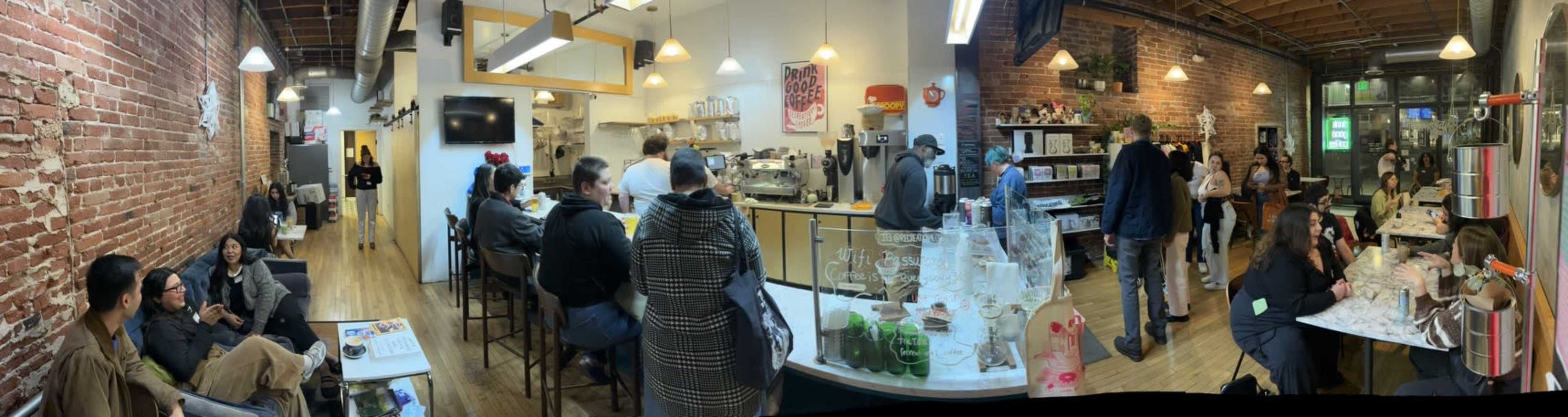 A spacious cafe features brick walls, a counter with coffee equipment, tables occupied by patrons, and a mix of seating arrangements.